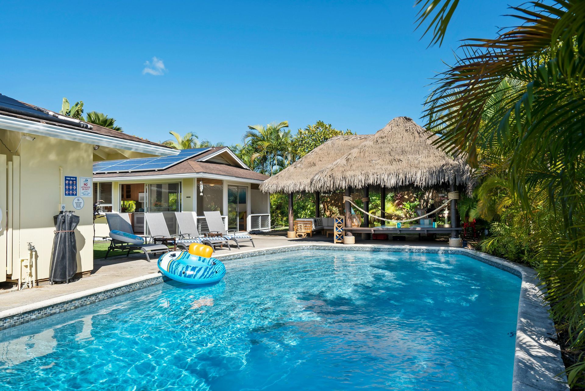 A tropical outdoor swimming pool with a blue inflatable float, lounge chairs, and a thatched-roof cabana under a clear sky.