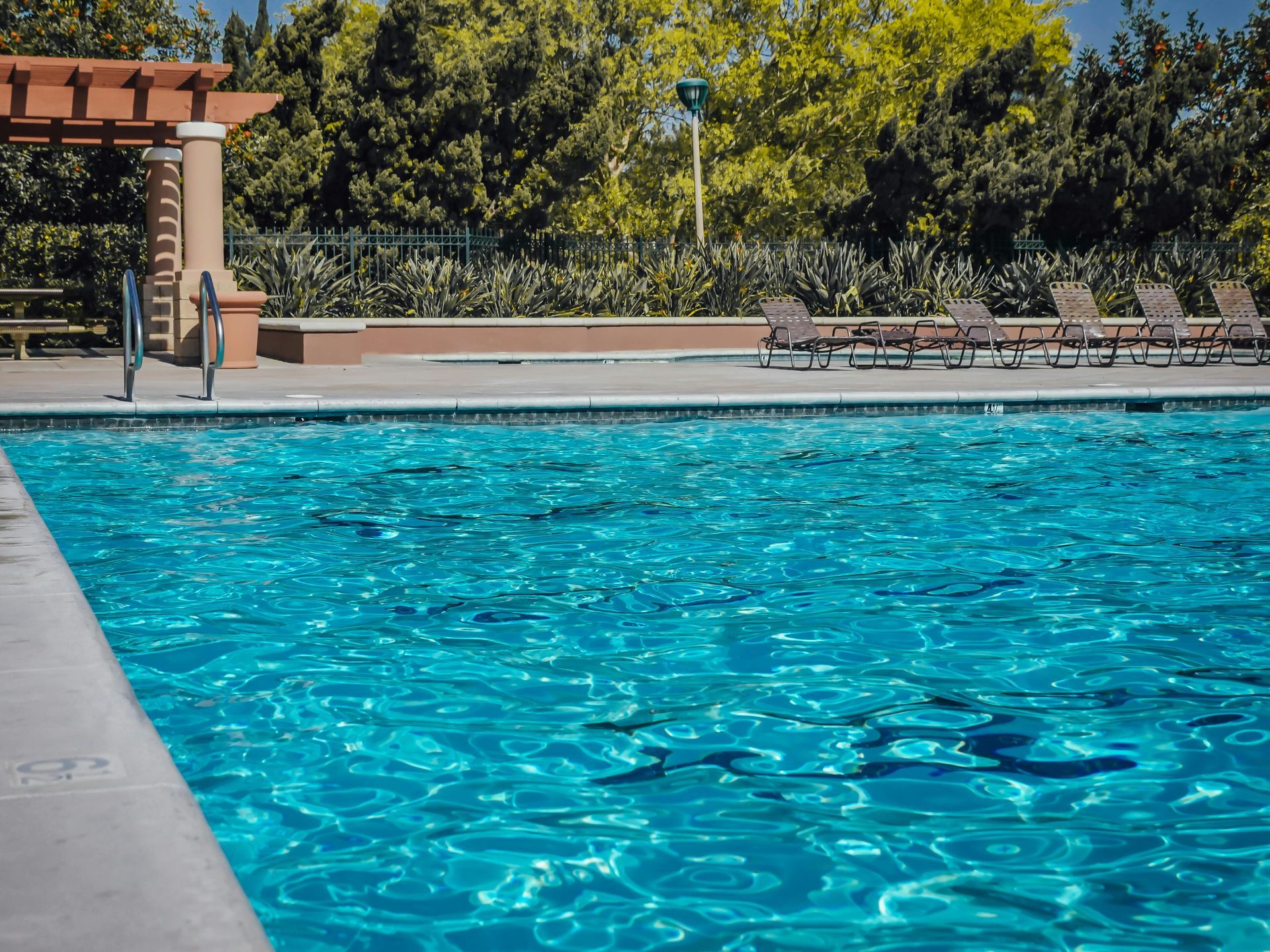 A sunny outdoor swimming pool with bright blue water, concrete decking, lounge chairs, and trees in the background.