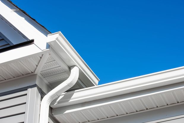 A white roof gutter and downspout installed on the corner of a house with light grey siding against a bright blue sky.