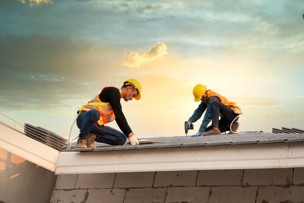 Two construction workers in yellow hard hats and orange safety vests install shingles on a roof at sunset.