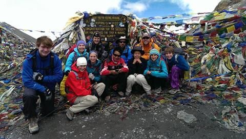 A group of hikers pose for a photo at a mountain pass summit covered in colorful prayer flags.