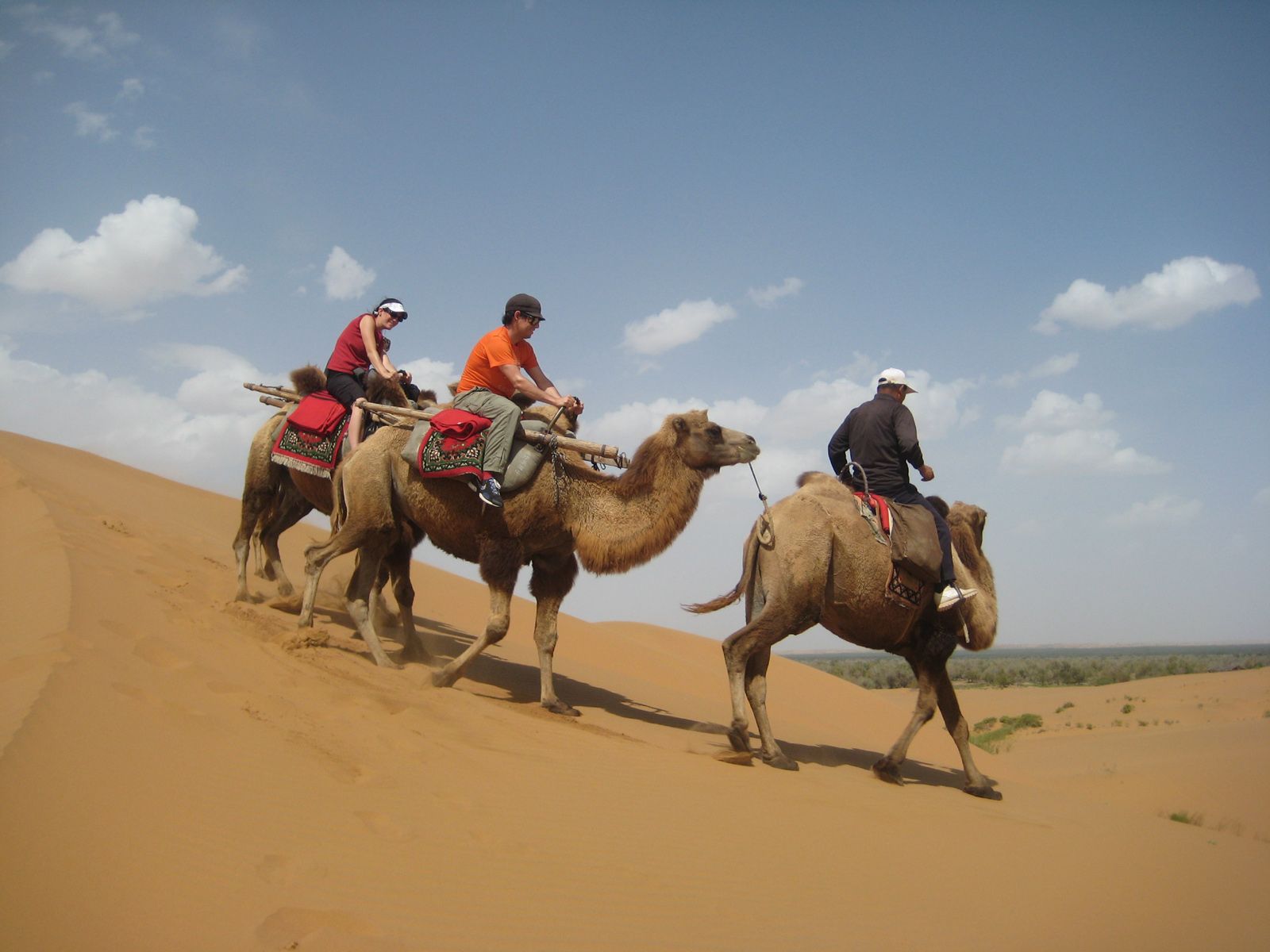 Three people ride camels across a large sand dune under a blue sky with scattered clouds.