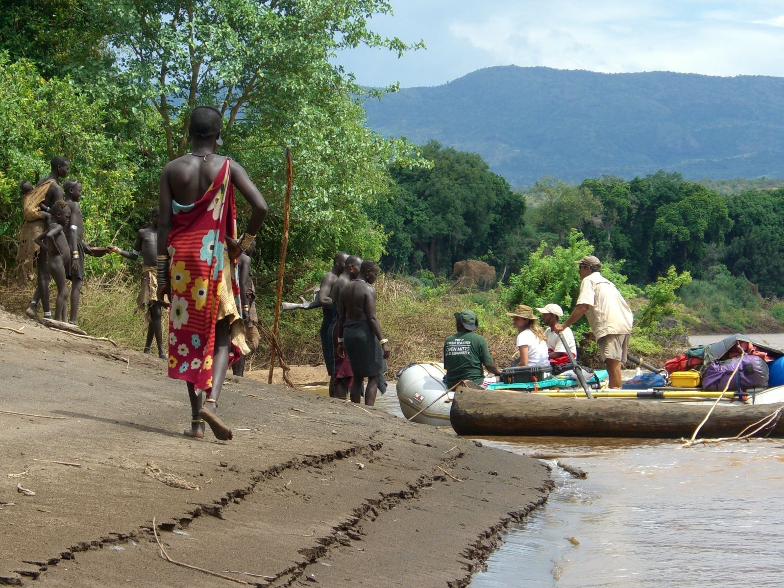 People in a raft arriving at a riverbank where individuals in patterned garments stand near a forest and distant mountains.
