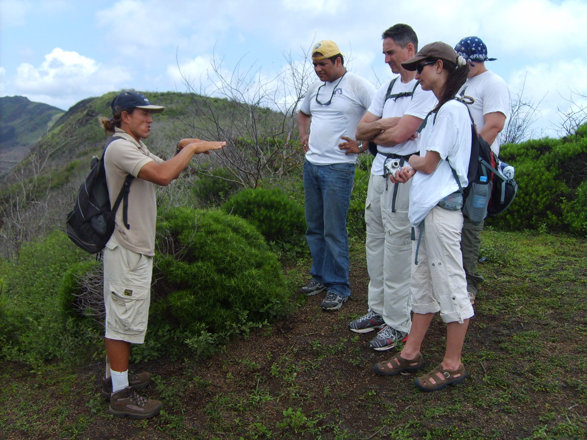 A guide gesturing to a group of hikers standing on a grassy, mountainous trail outdoors.