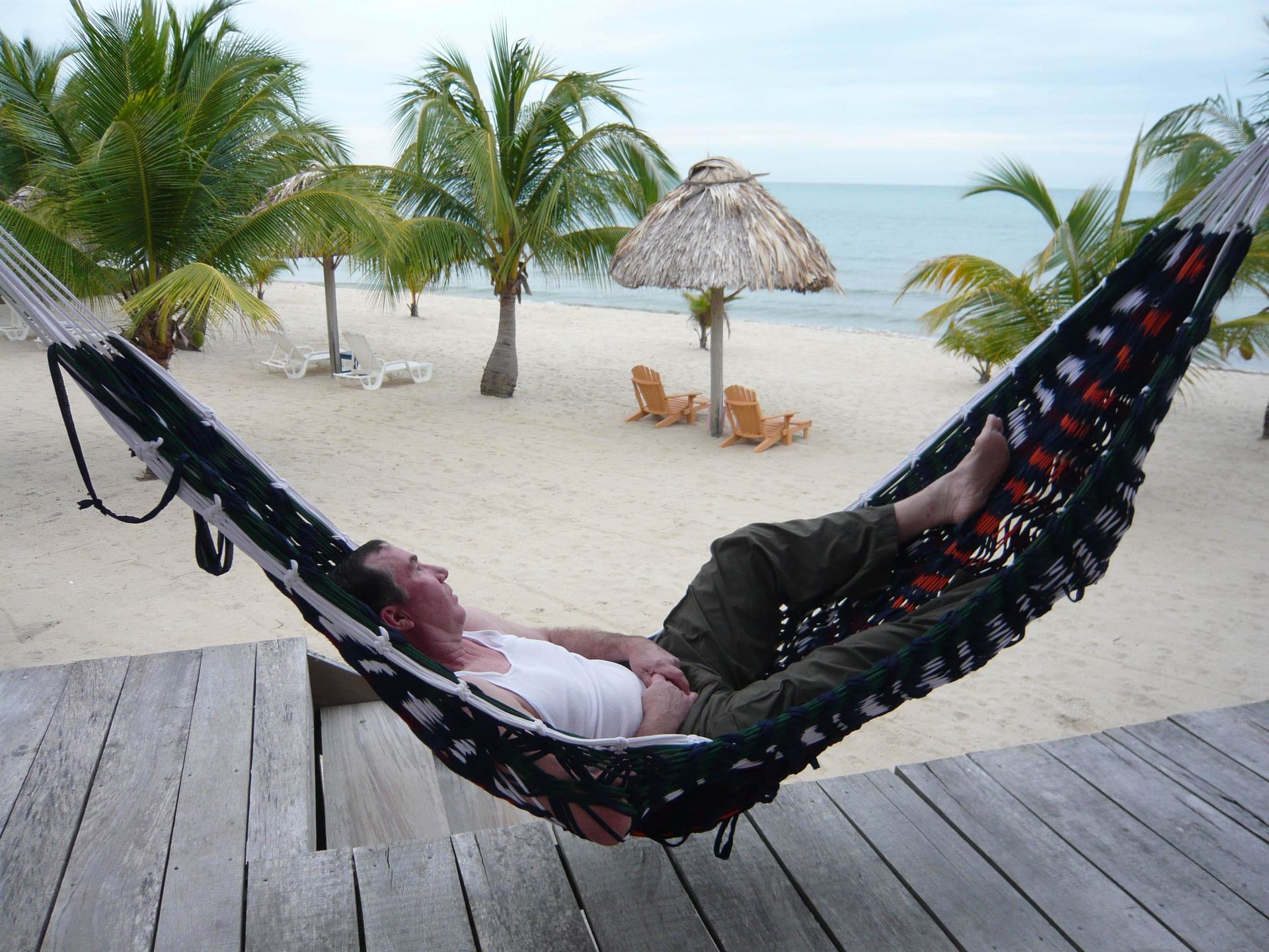 A person rests in a black hammock on a wooden deck overlooking a sandy beach with palm trees and a thatched umbrella.