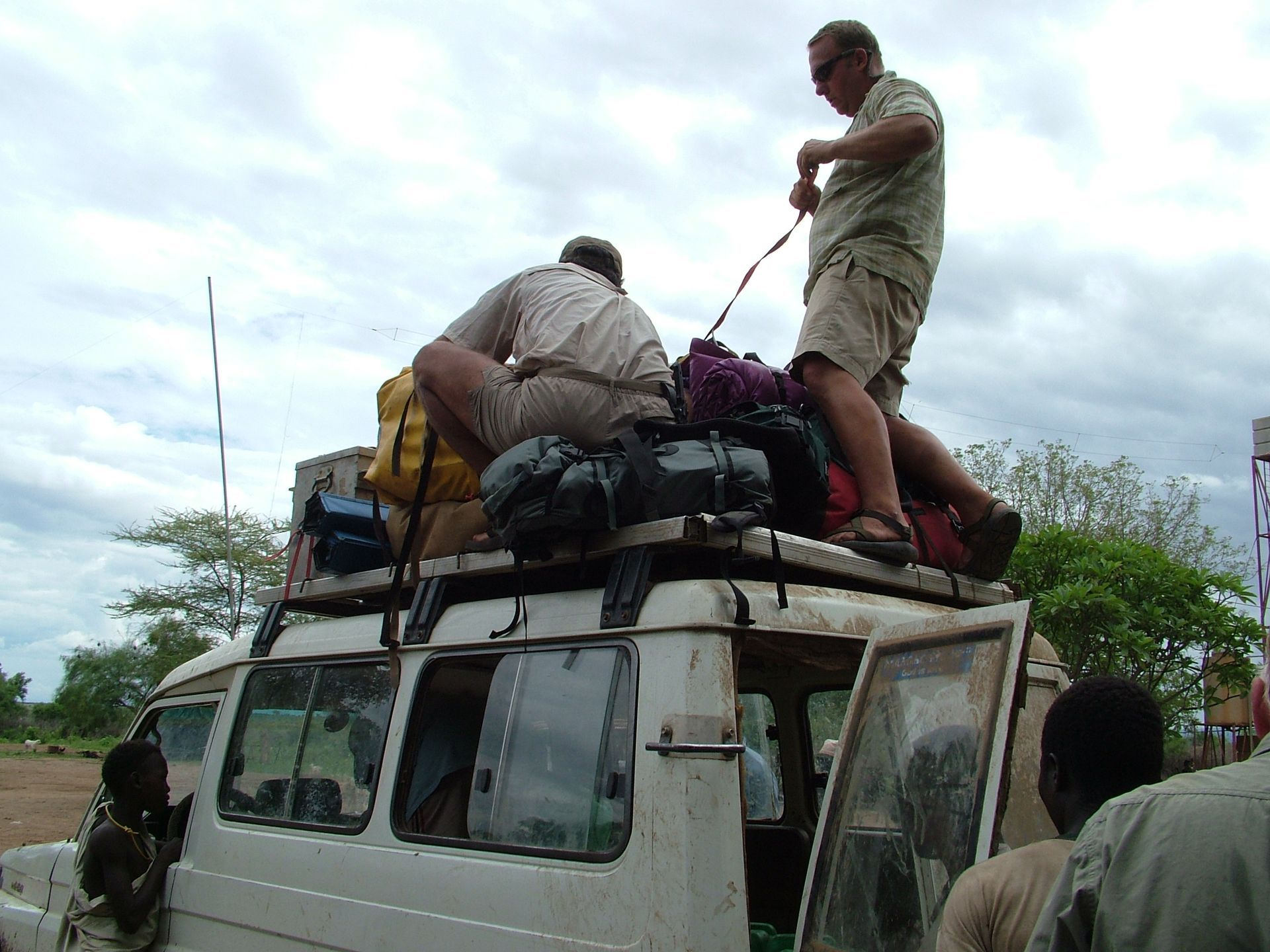 Two people secure a large pile of luggage and backpacks onto the roof rack of a white van parked outdoors.