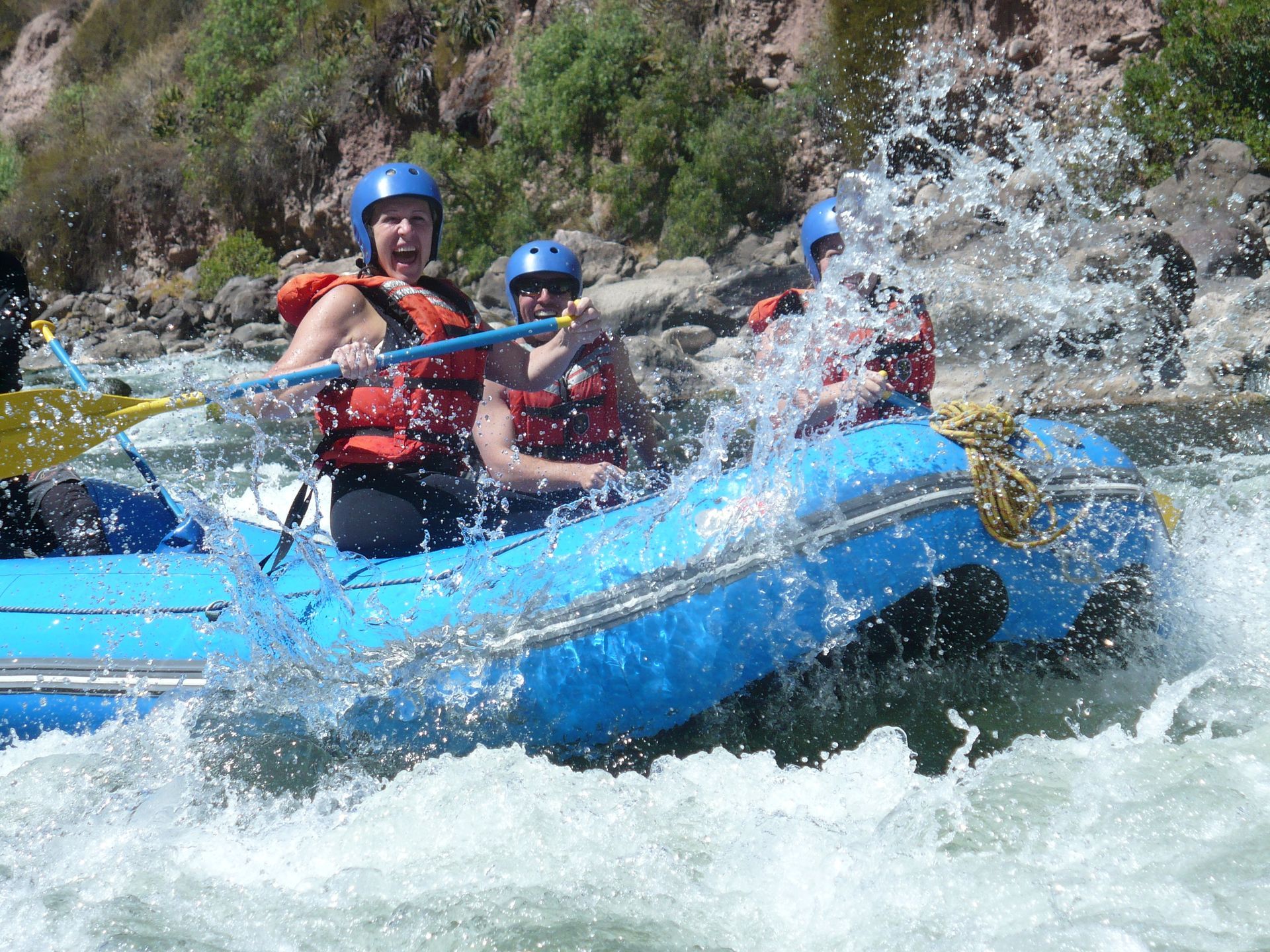 People in helmets and life vests paddling a bright blue raft through splashing river rapids.