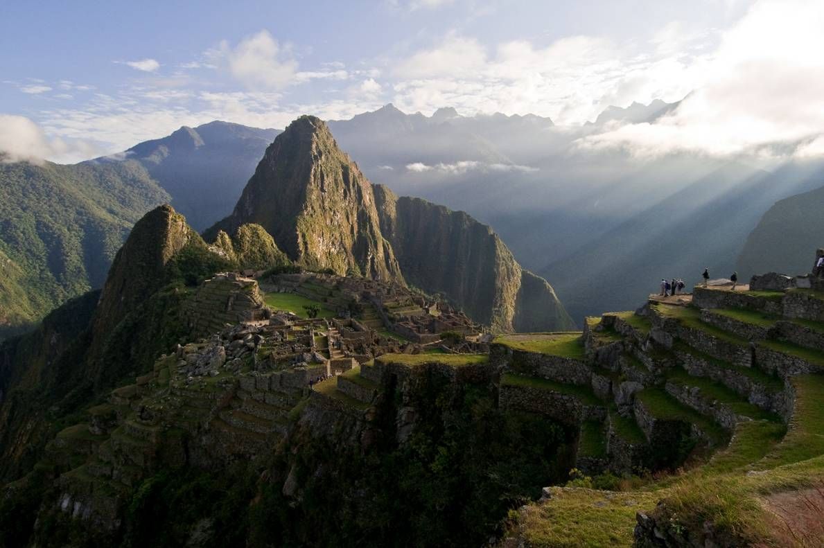 The ruins of Machu Picchu with stone terraces in the foreground and a large mountain peak in the background at sunrise.