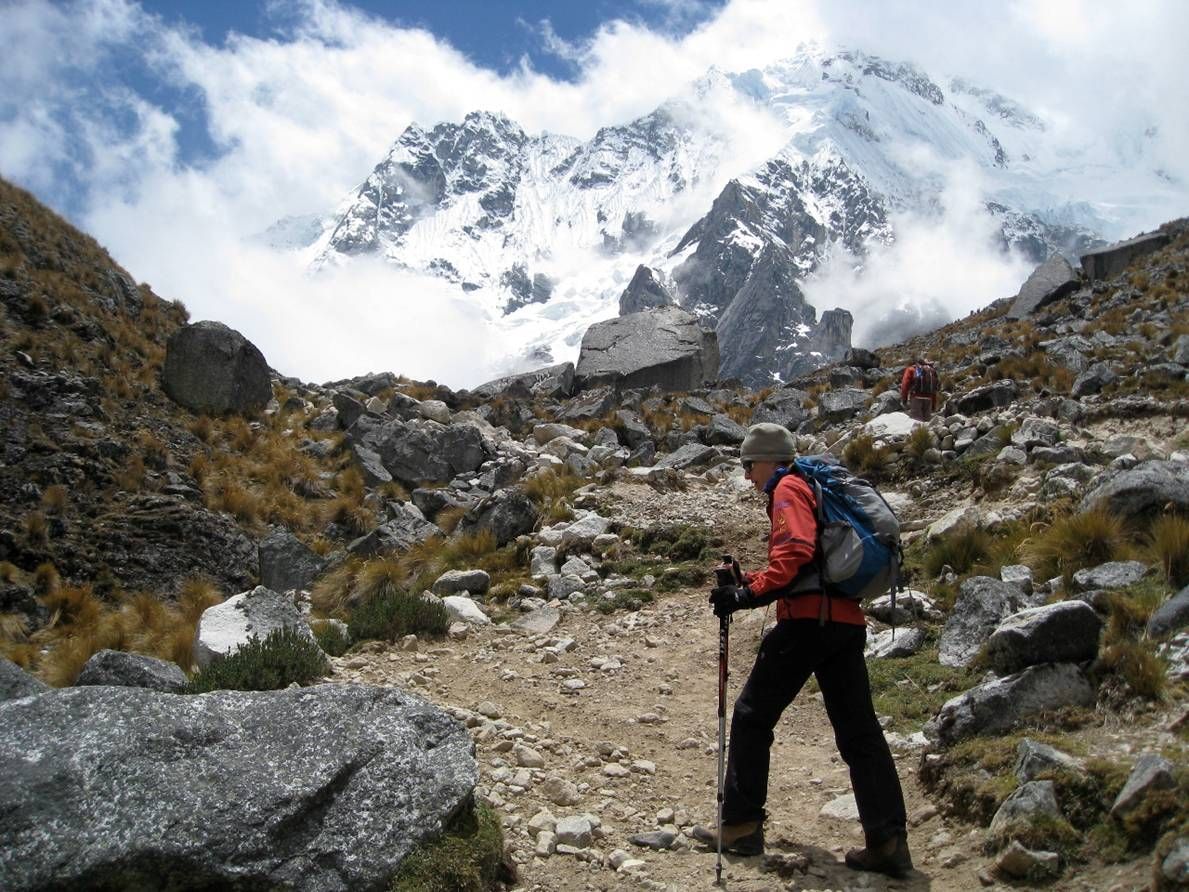 A hiker with a backpack and trekking poles walks up a rocky trail toward a snow-capped mountain under a cloudy sky.