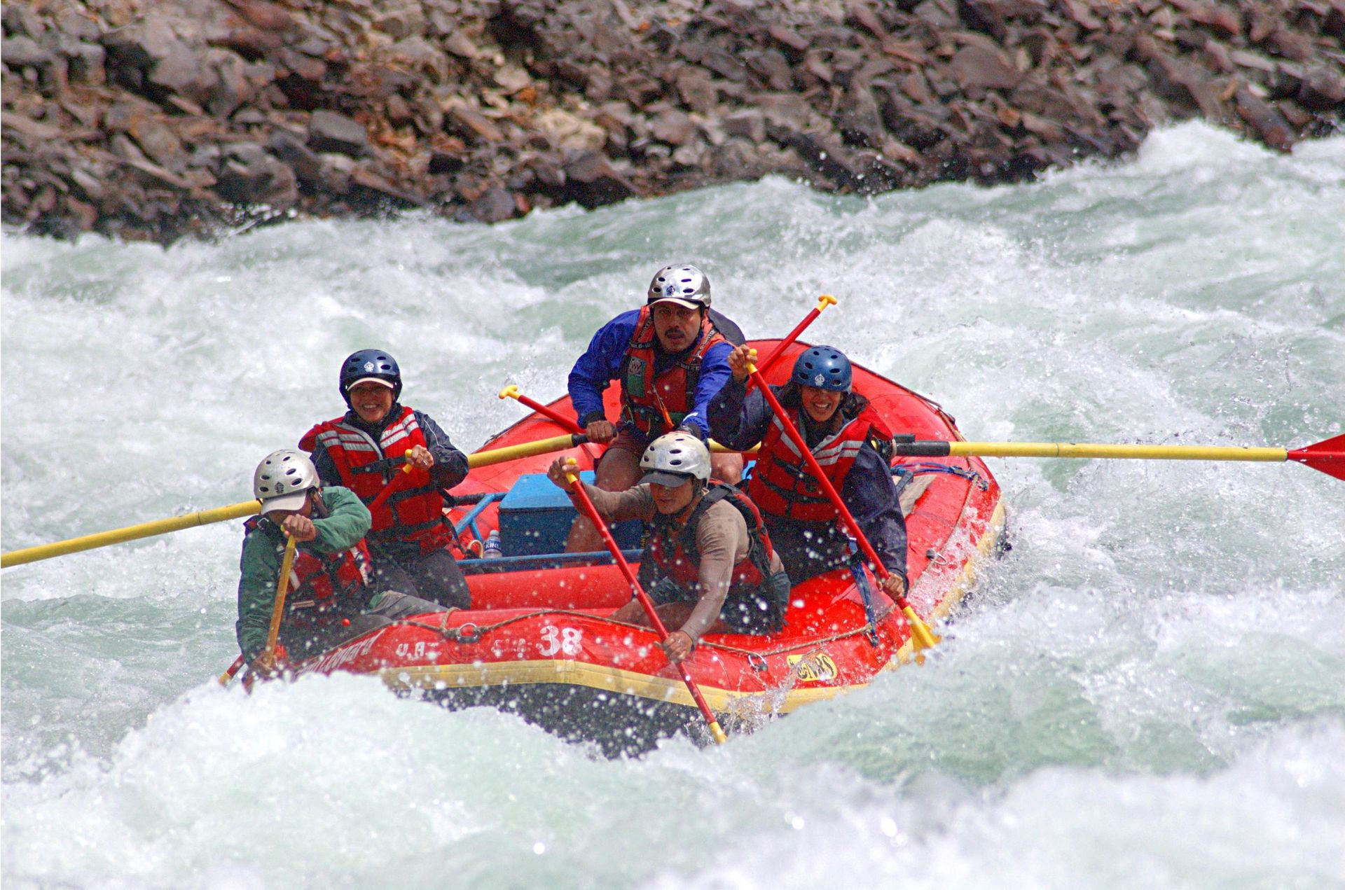 Four people wearing life jackets and helmets paddle a red raft through white-water rapids next to a rocky bank.