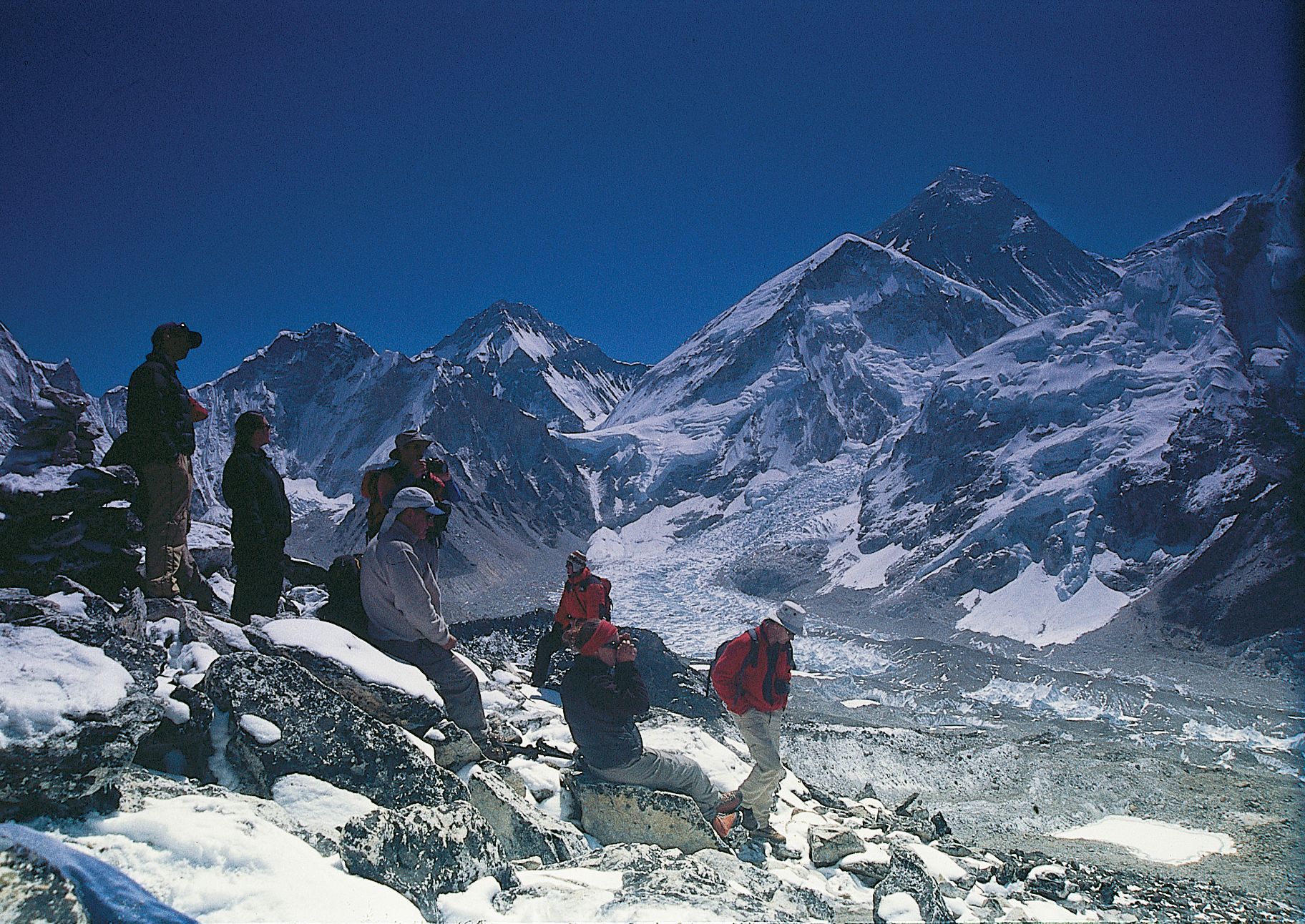 A group of hikers in outdoor gear pauses on a rocky, snow-dusted ridge looking out toward a massive, snow-covered mountain.