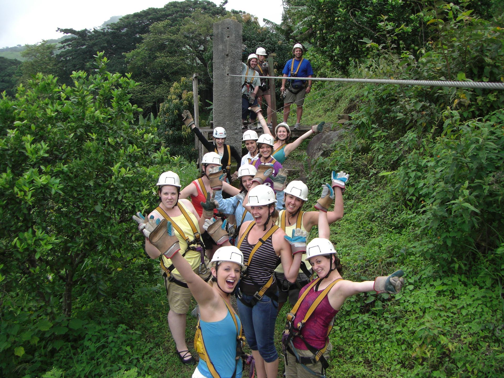 A group wearing climbing harnesses and helmets poses happily on a zip-line platform amidst a lush, wooded area.