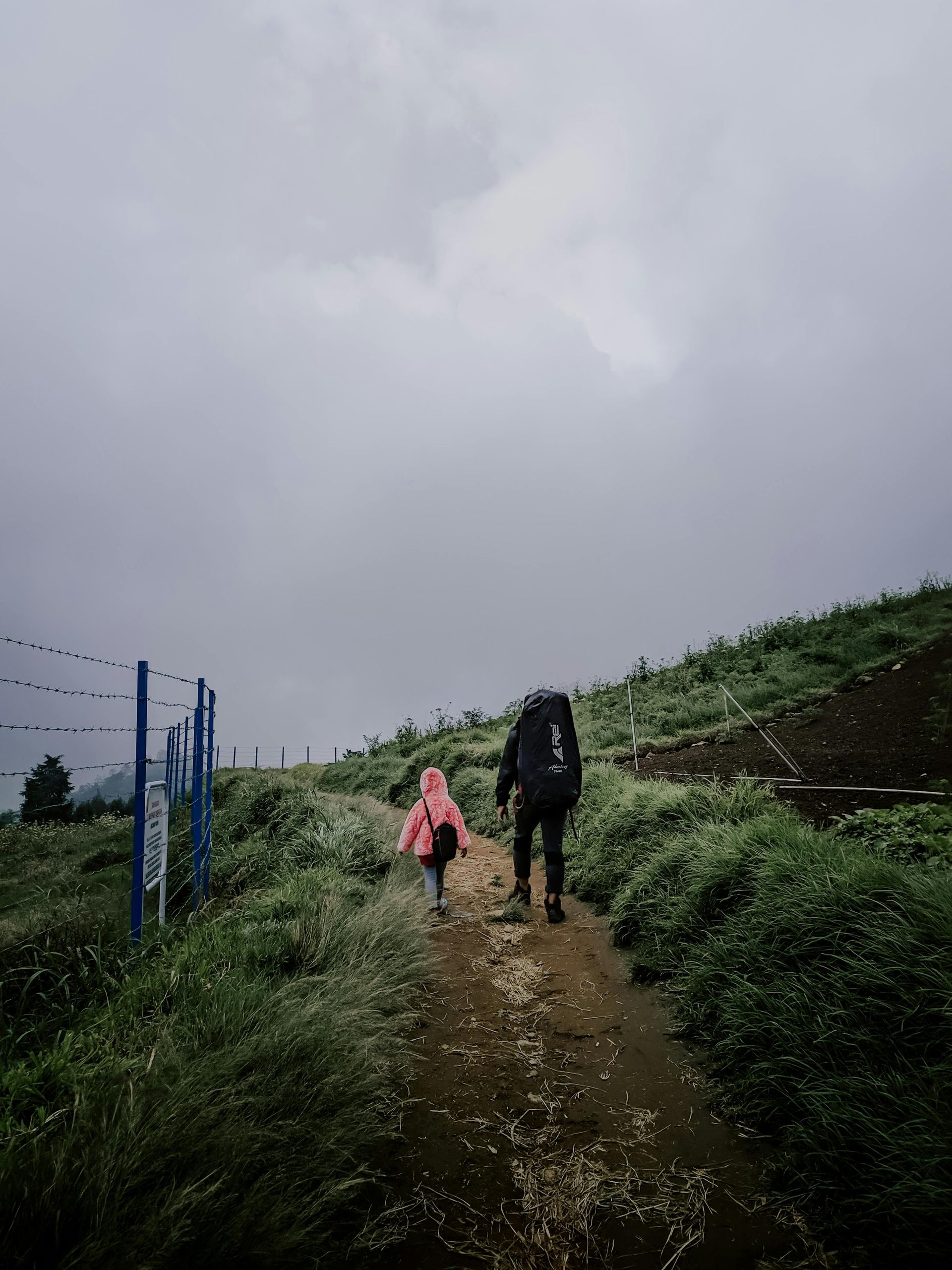 A person with a large backpack walks alongside a smaller person on a dirt path under a cloudy sky near a wire fence.