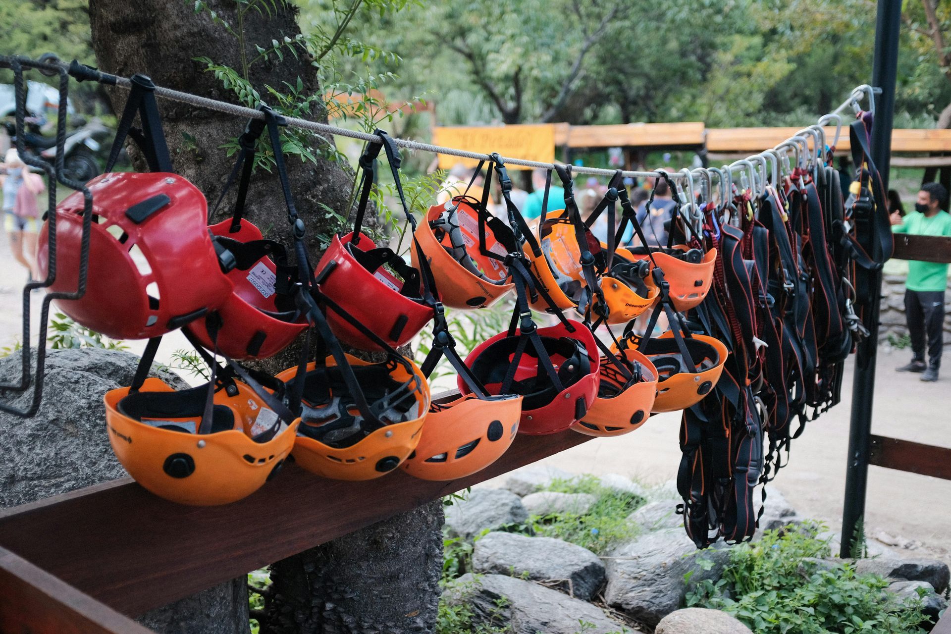 Rows of red and orange climbing helmets and harnesses hanging from a rope outdoors at an adventure park.
