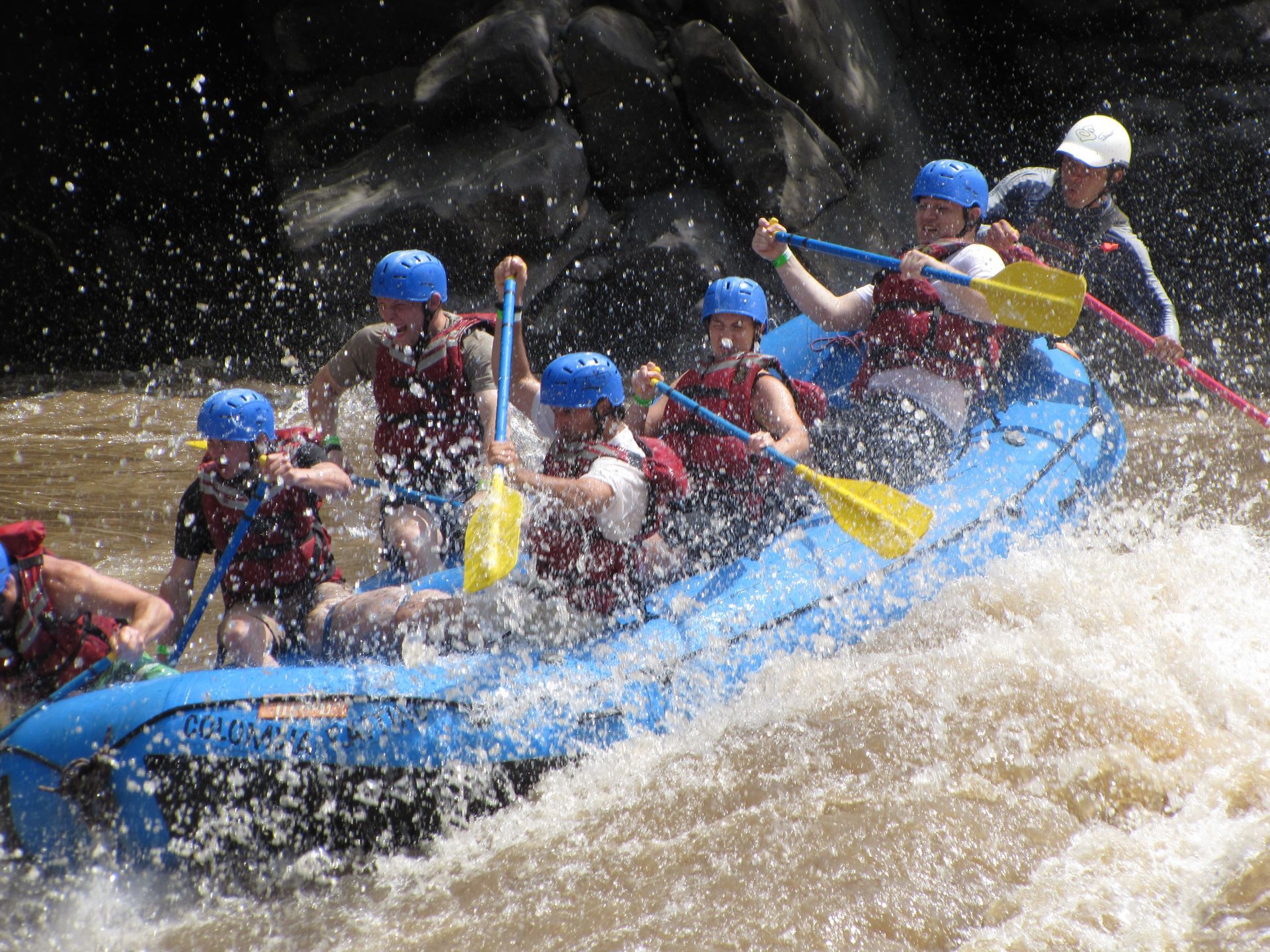 A group of people wearing helmets and life vests paddling a blue raft through splashing whitewater rapids.