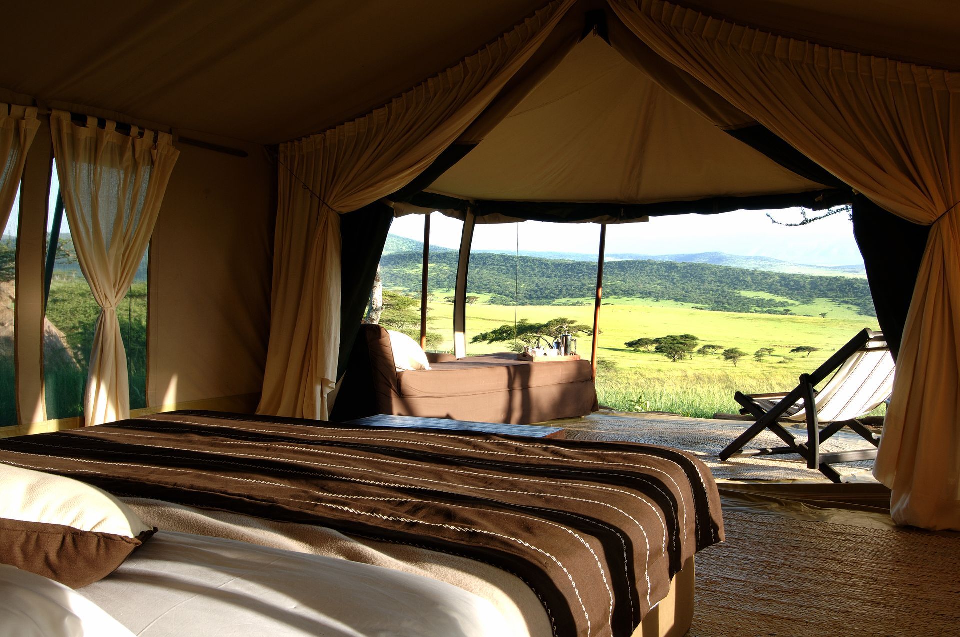 A luxurious safari tent bedroom with an open front overlooking a lush, sunlit savanna landscape.