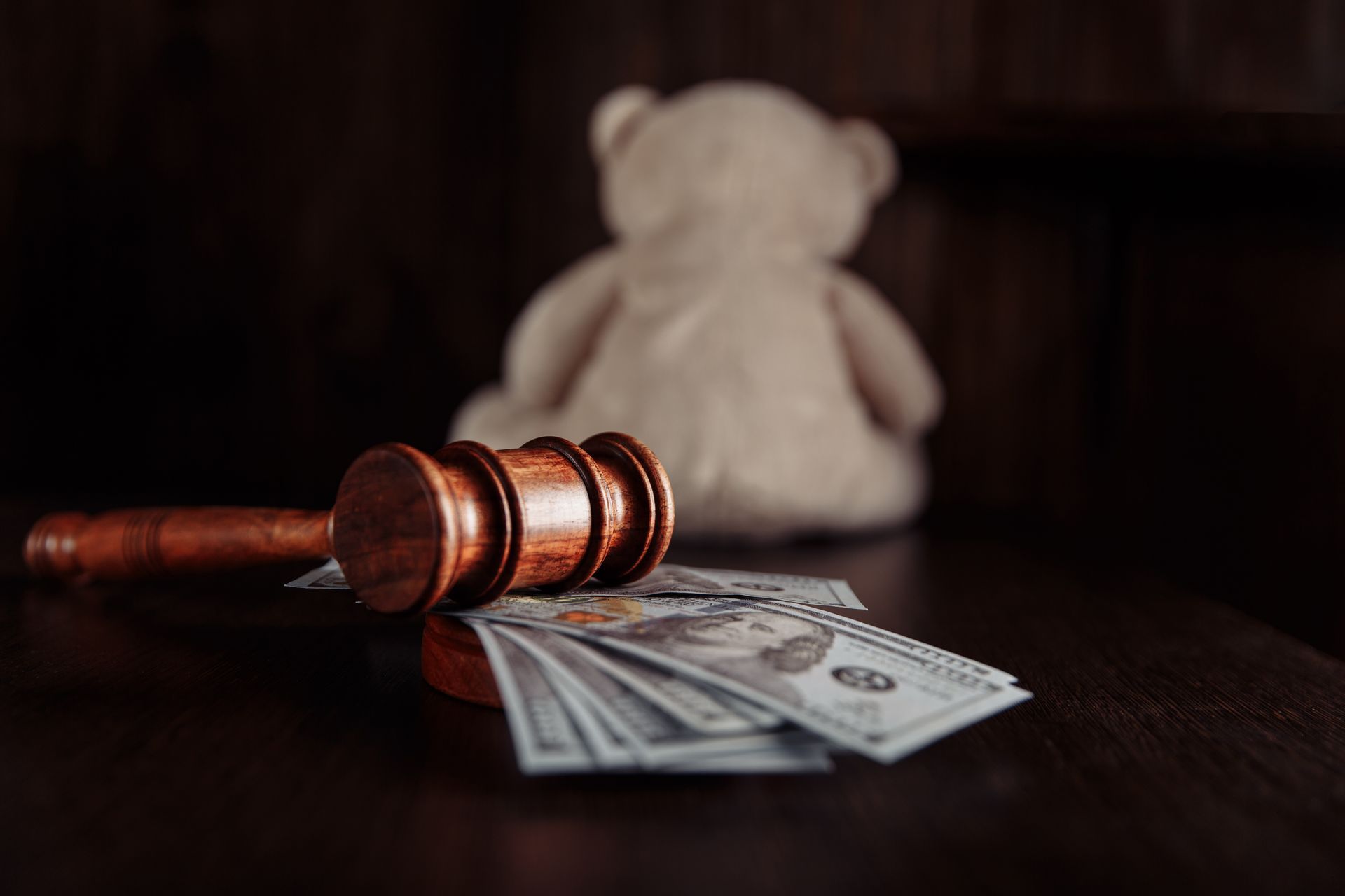 Gavel, cash, and teddy bear on a dark table, possibly a court setting.