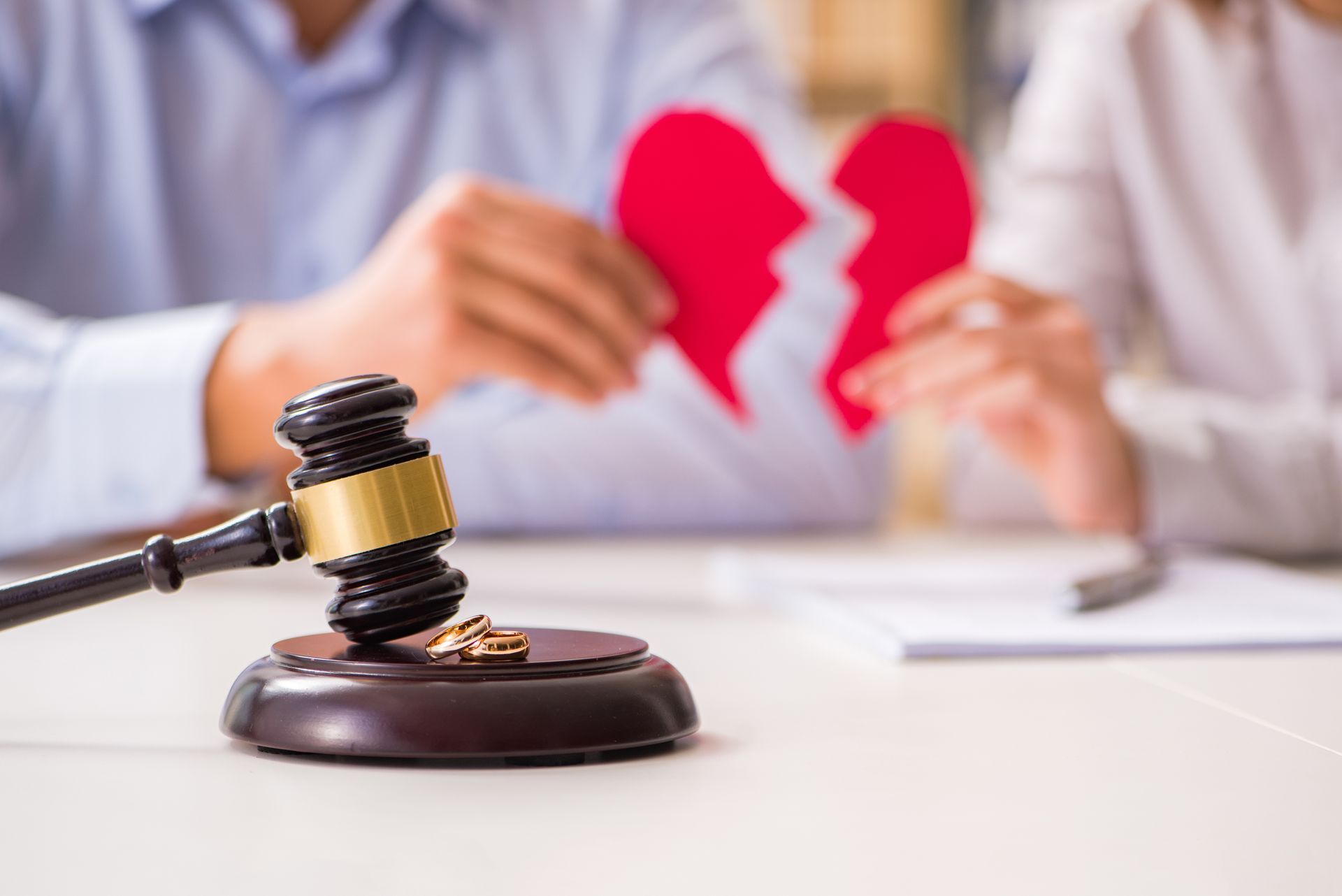 Gavel, broken heart, and wedding rings on a table; a couple in the background.