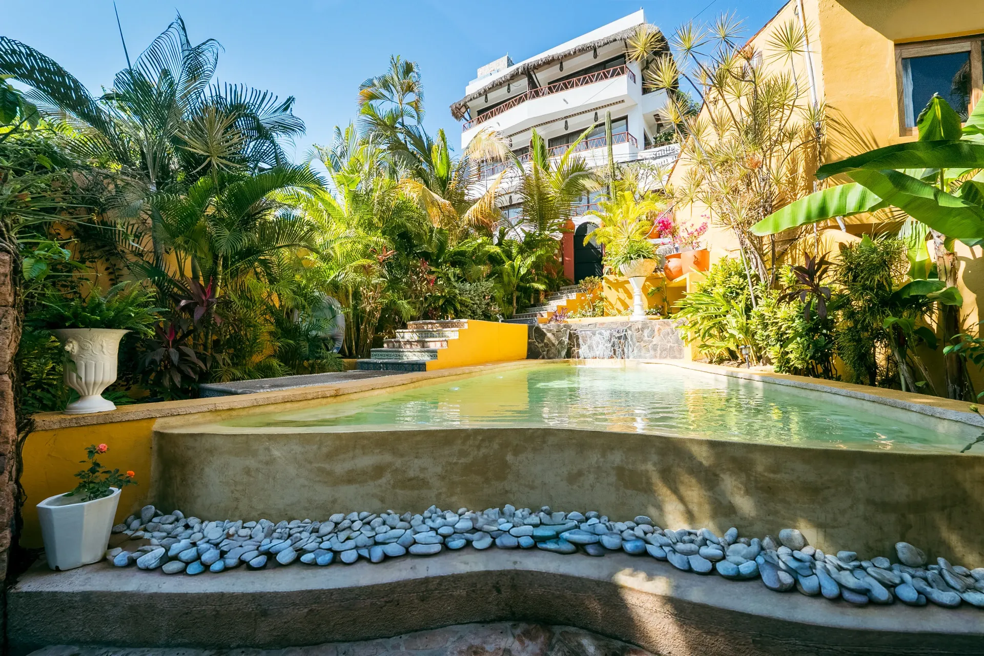 Pool surrounded by lush greenery and a multi-story building. Water flows over a small waterfall.