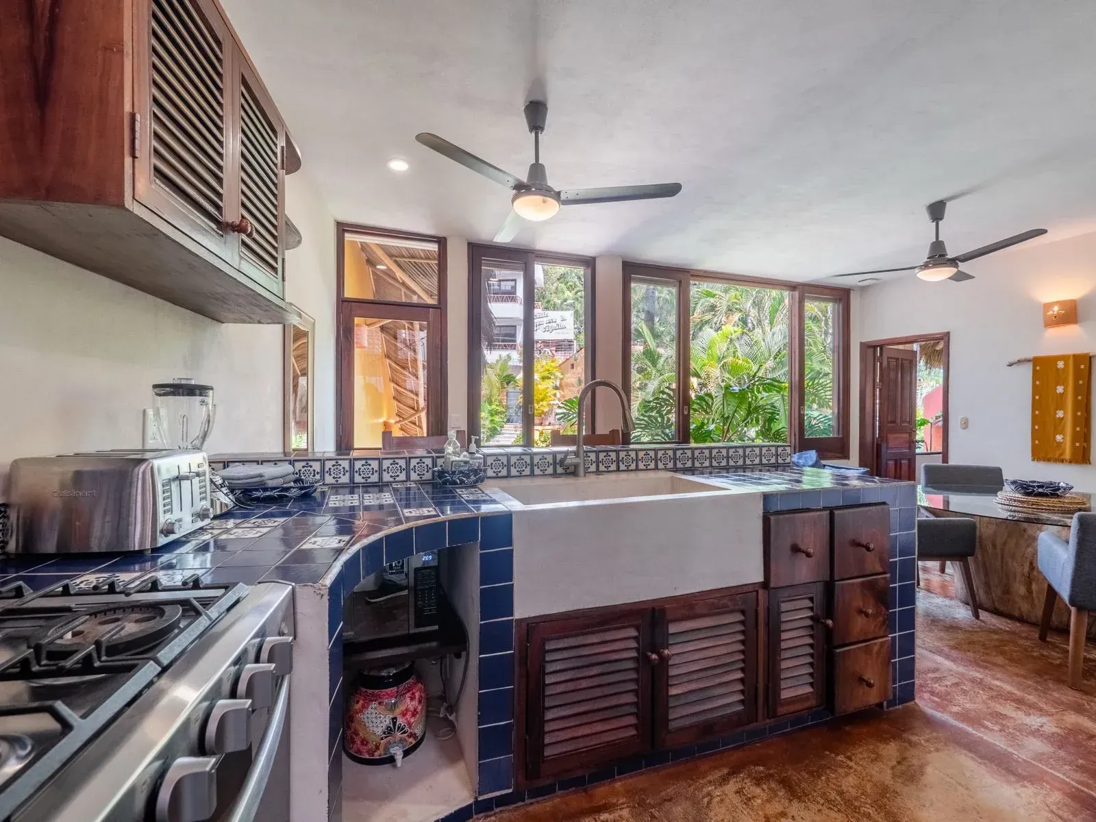 Kitchen with blue tiled countertops, farmhouse sink, wooden cabinets, and large windows overlooking greenery.