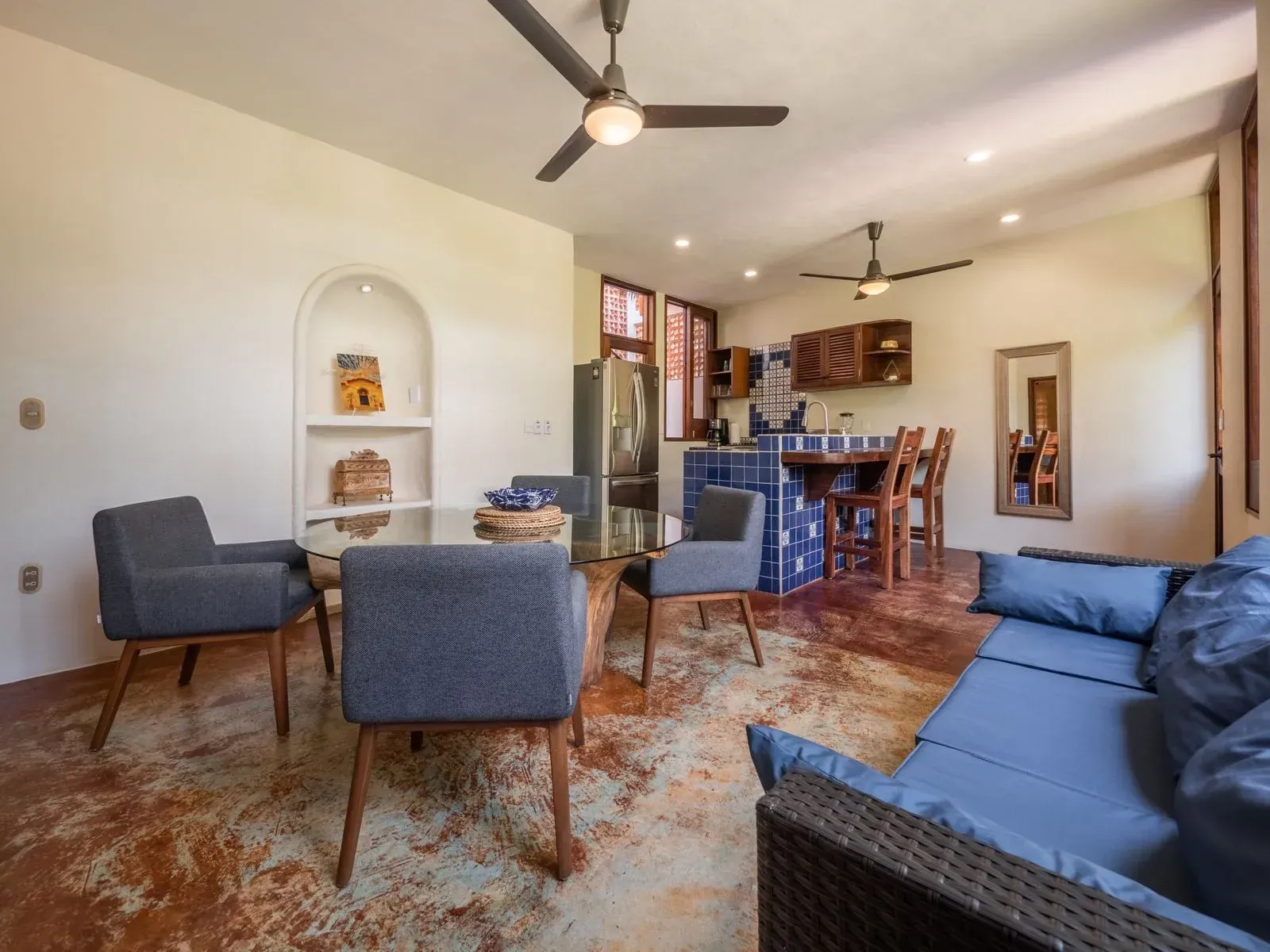 An open living space with a dining area, kitchen and blue couch. The walls are white and the floors are brown.