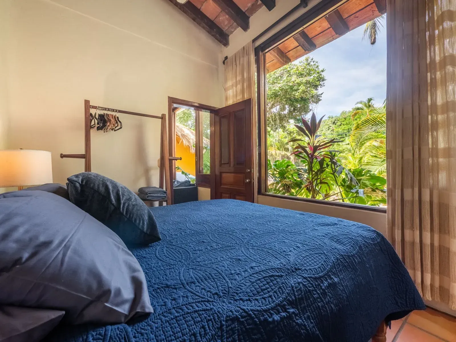 Bedroom with blue bedspread, open window showing lush greenery, wooden rack for clothes.