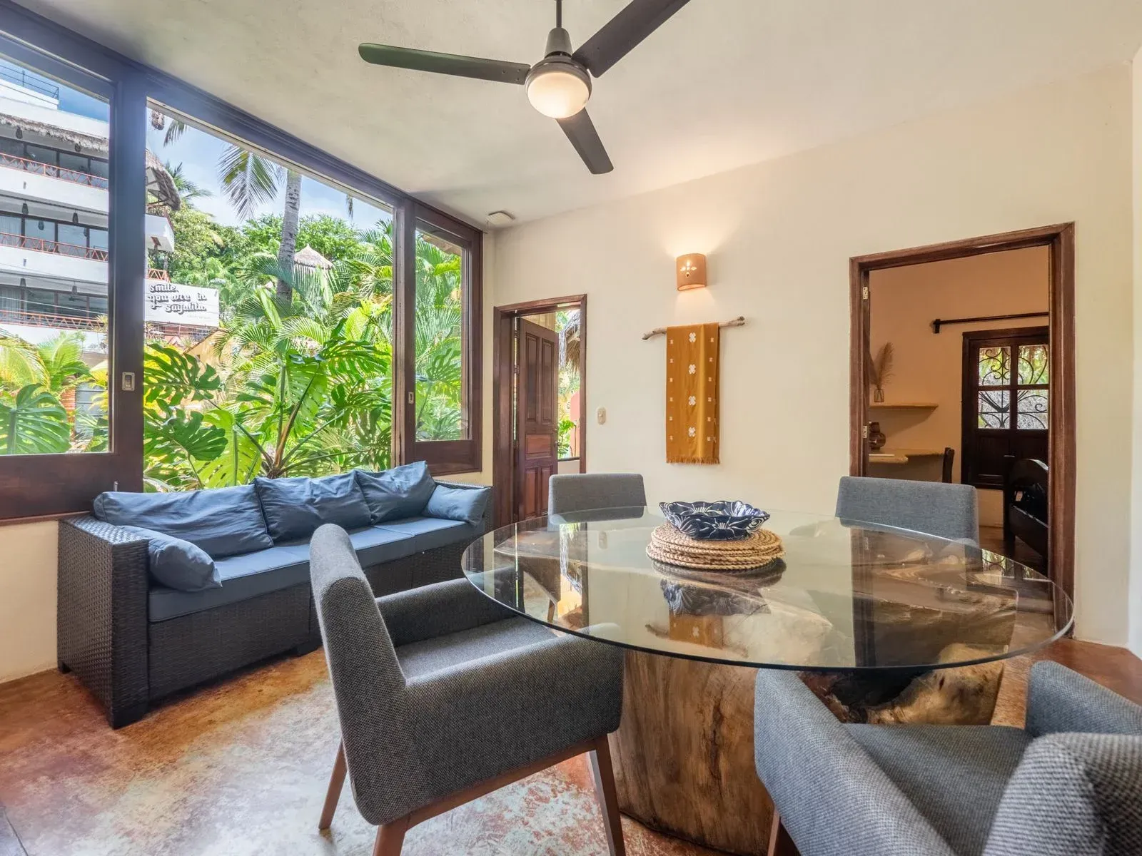Dining area with glass table, gray chairs, and a dark blue sofa next to a window overlooking greenery.