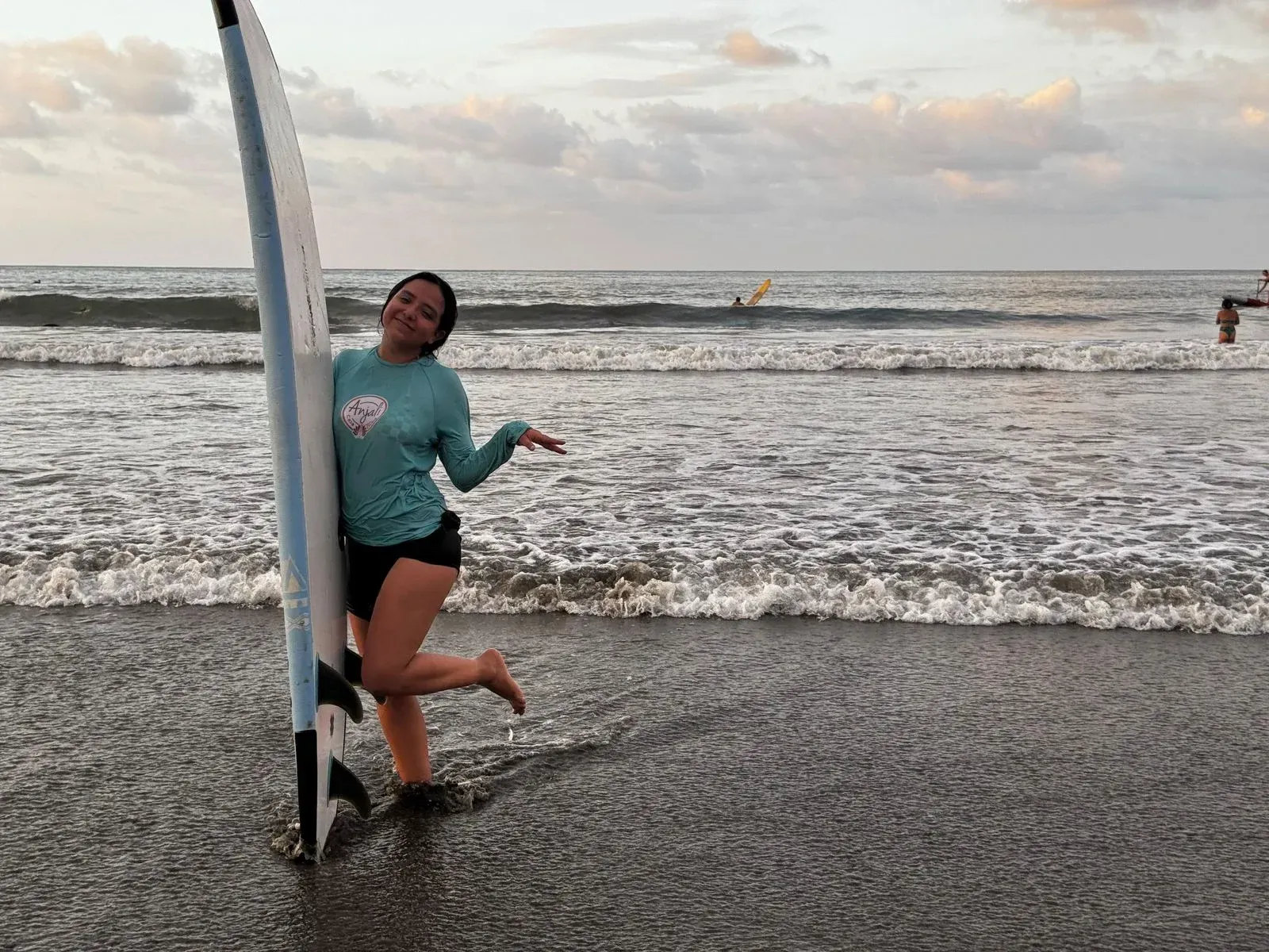 Woman with surfboard smiles at beach, waves gently lap at her feet.