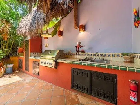 Outdoor kitchen with grill, sink, and storage beneath a thatched roof. Orange, teal, and beige.