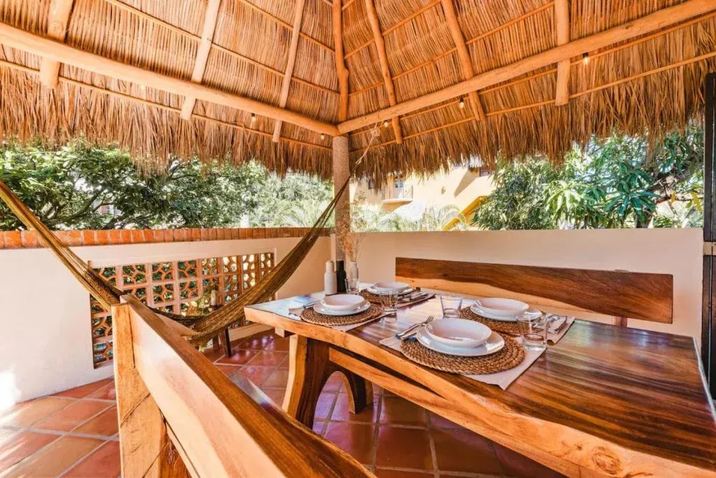 Outdoor dining area with thatched roof, wooden table and bench, hammock, and tiled floor.