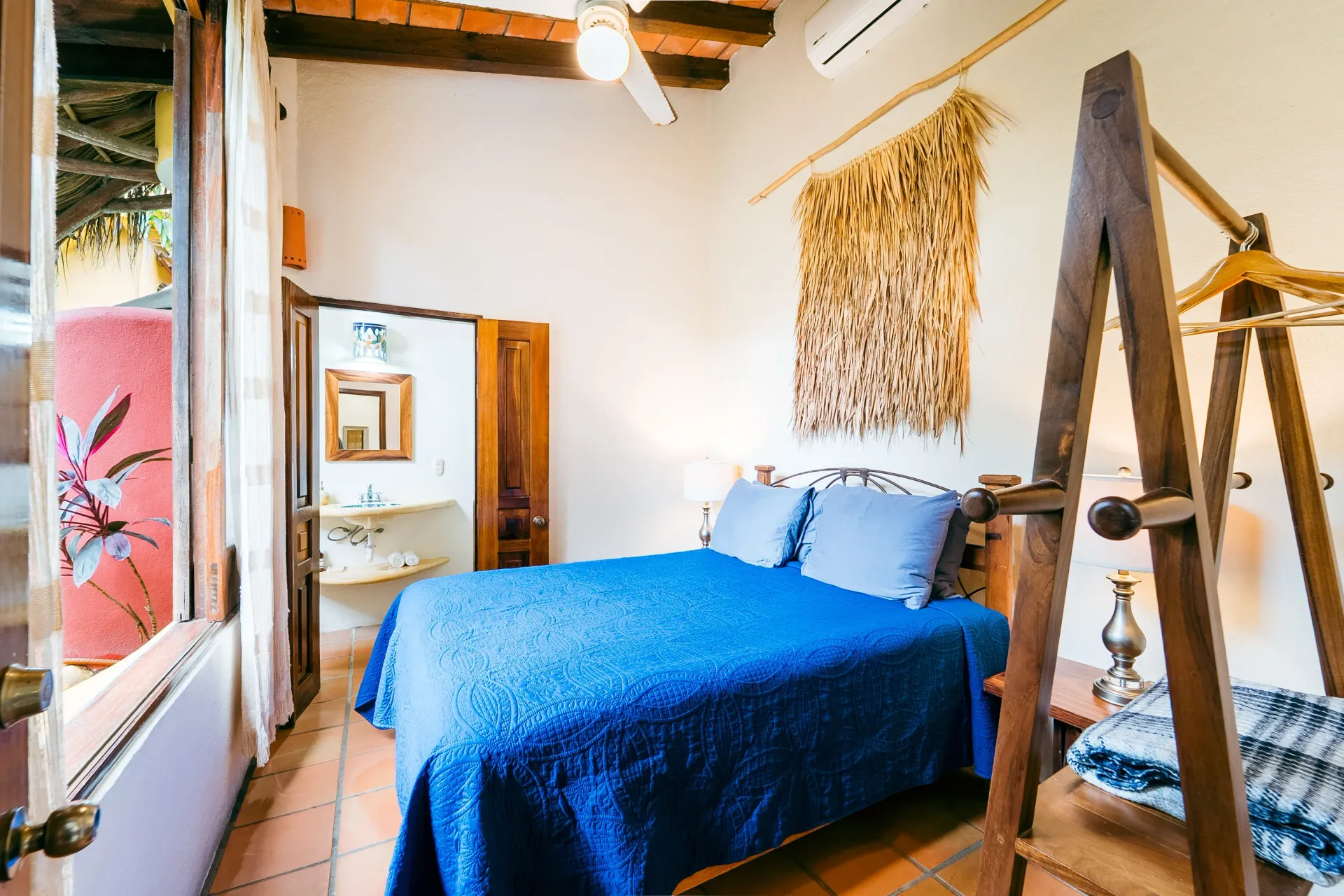 Bedroom with blue bed, open doorway to bathroom, clay tile floor. Rustic decor.