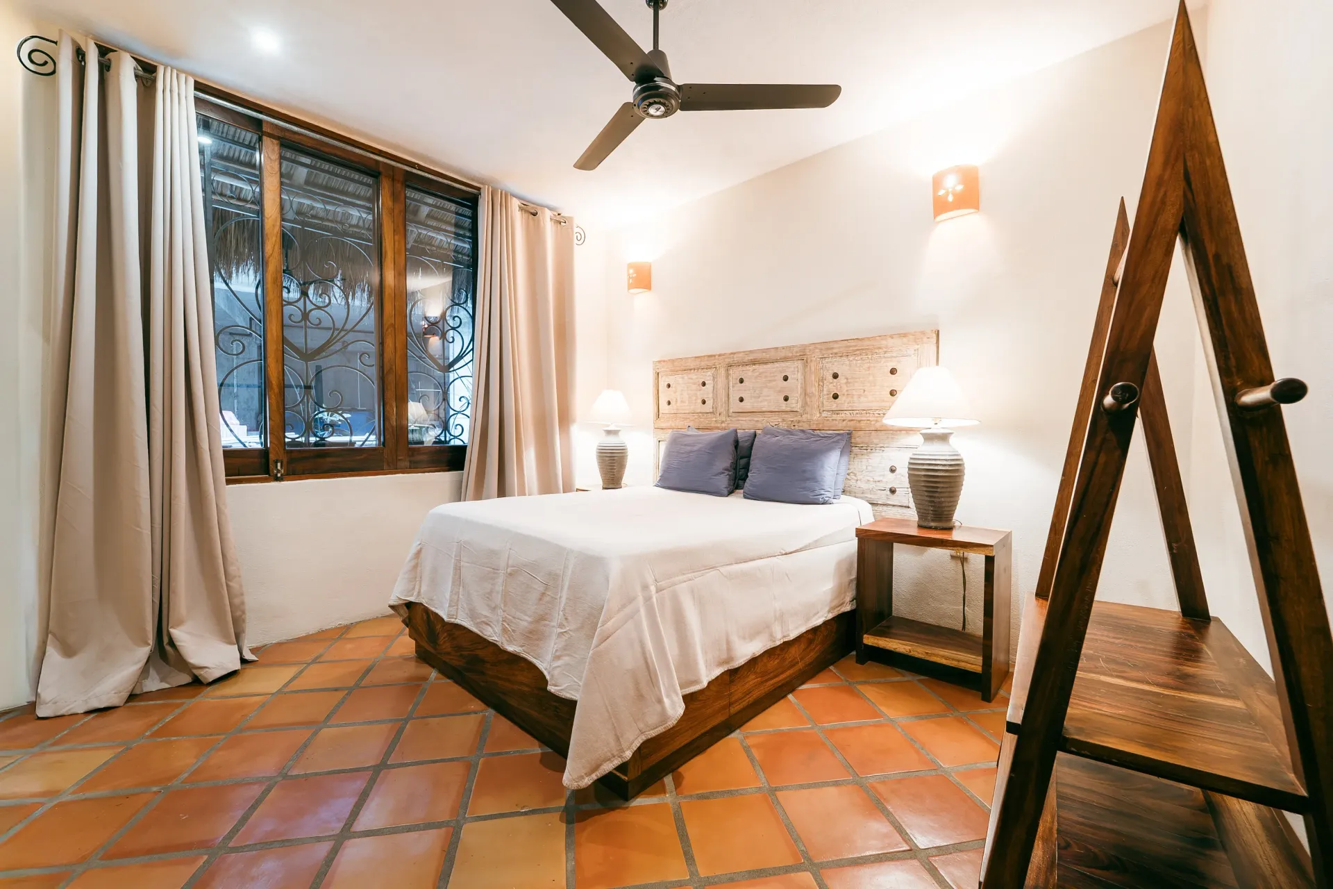 Bedroom with a bed, nightstand, and window. Terra-cotta tile floor, off-white walls, and a ceiling fan.