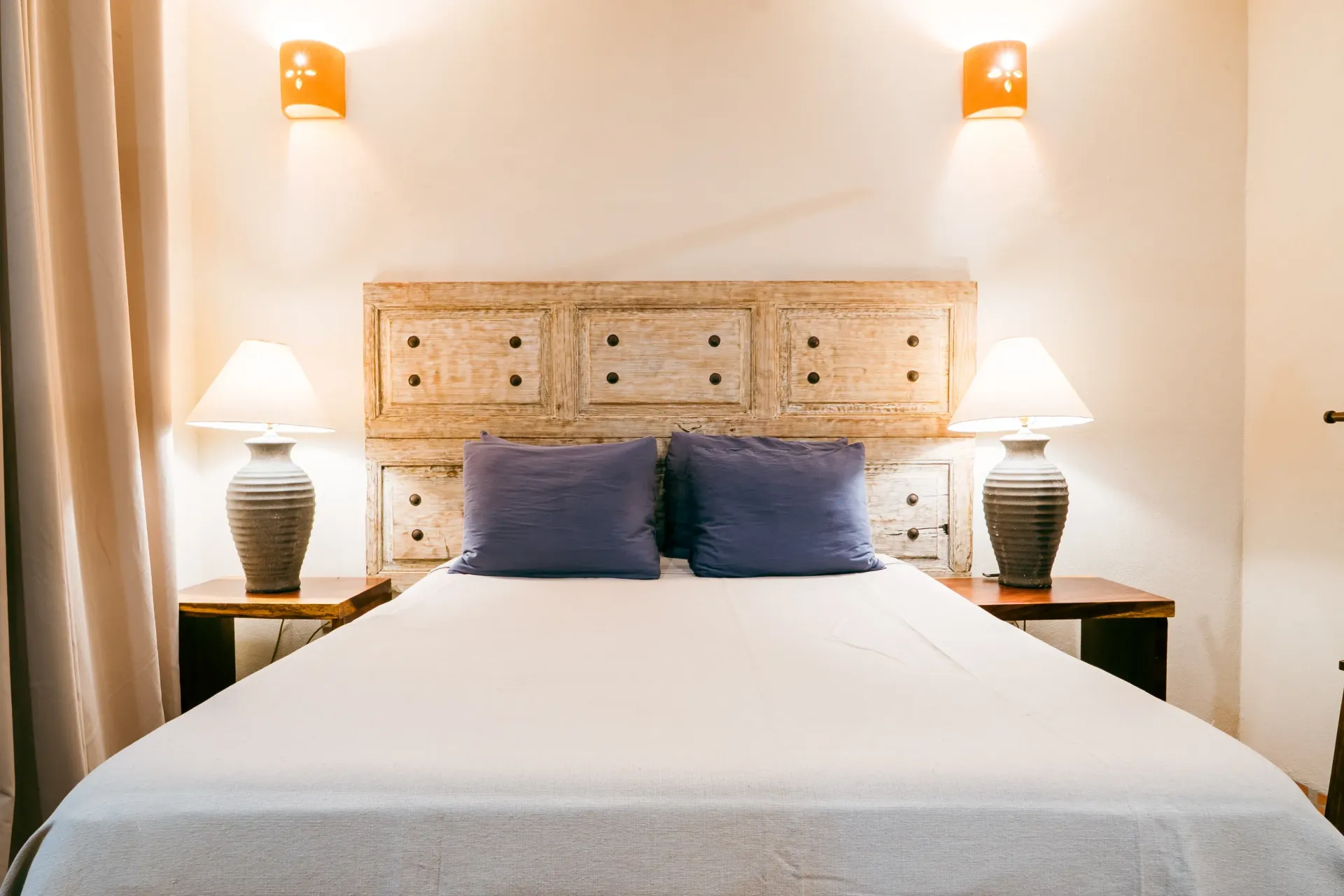 Bedroom with a white bed, blue pillows, wooden headboard, lamps on nightstands, and wall sconces.