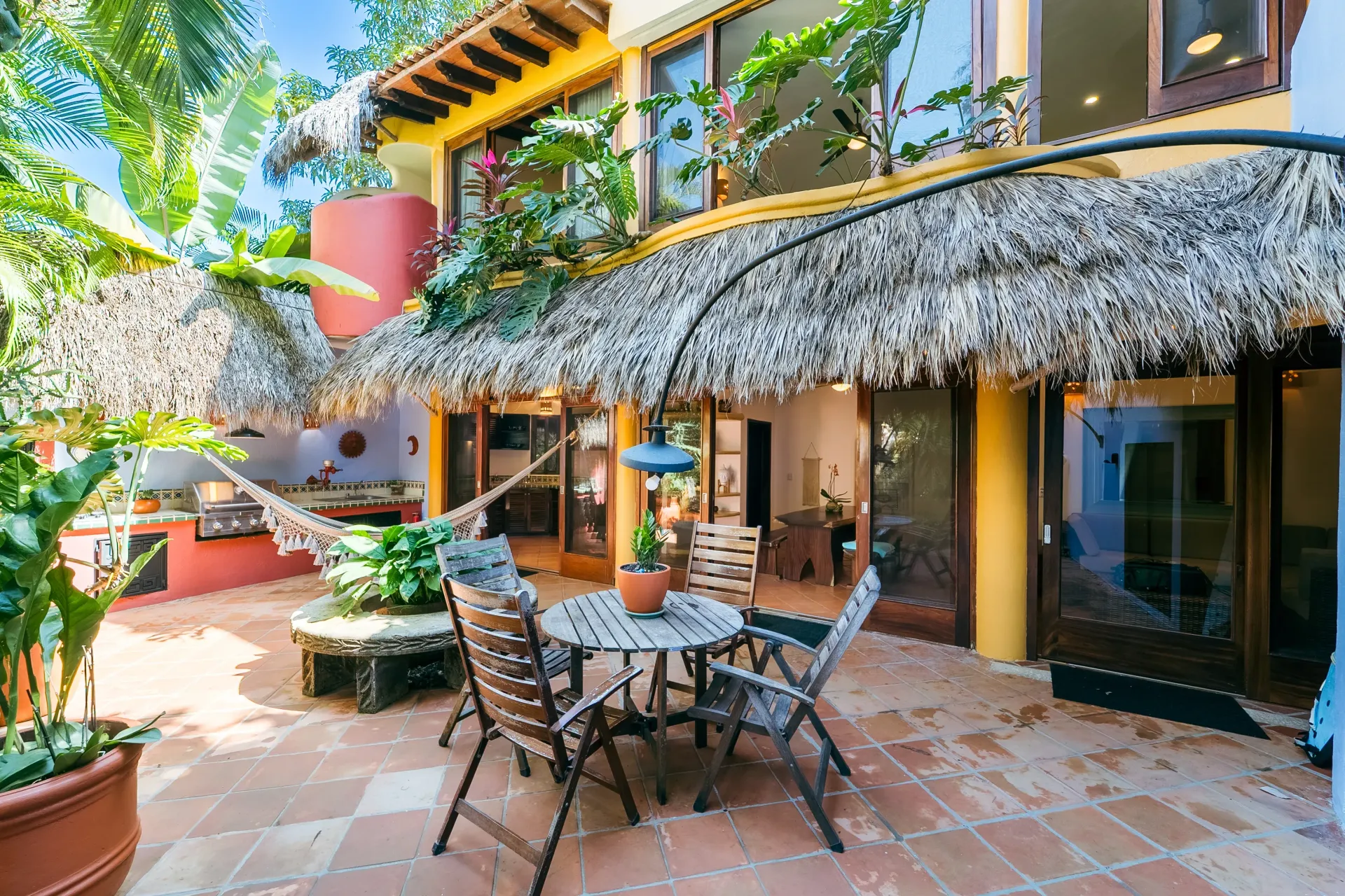 Patio with thatched roof, dining table, hammock, potted plants, and two-story building in the background.