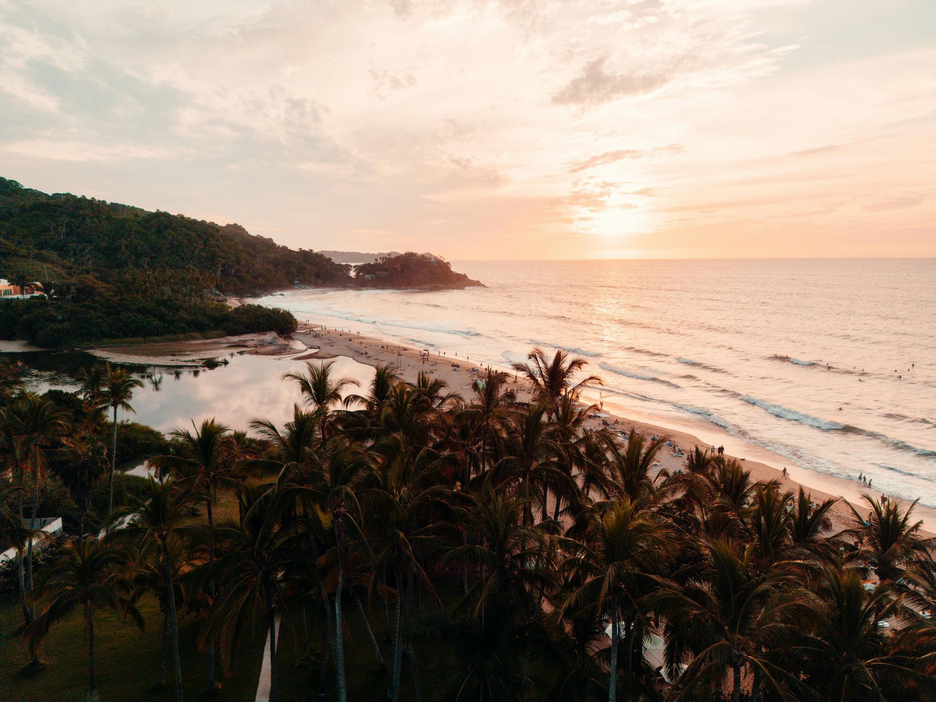 Palm trees frame a beach at sunset, with waves rolling in and people on the shore.