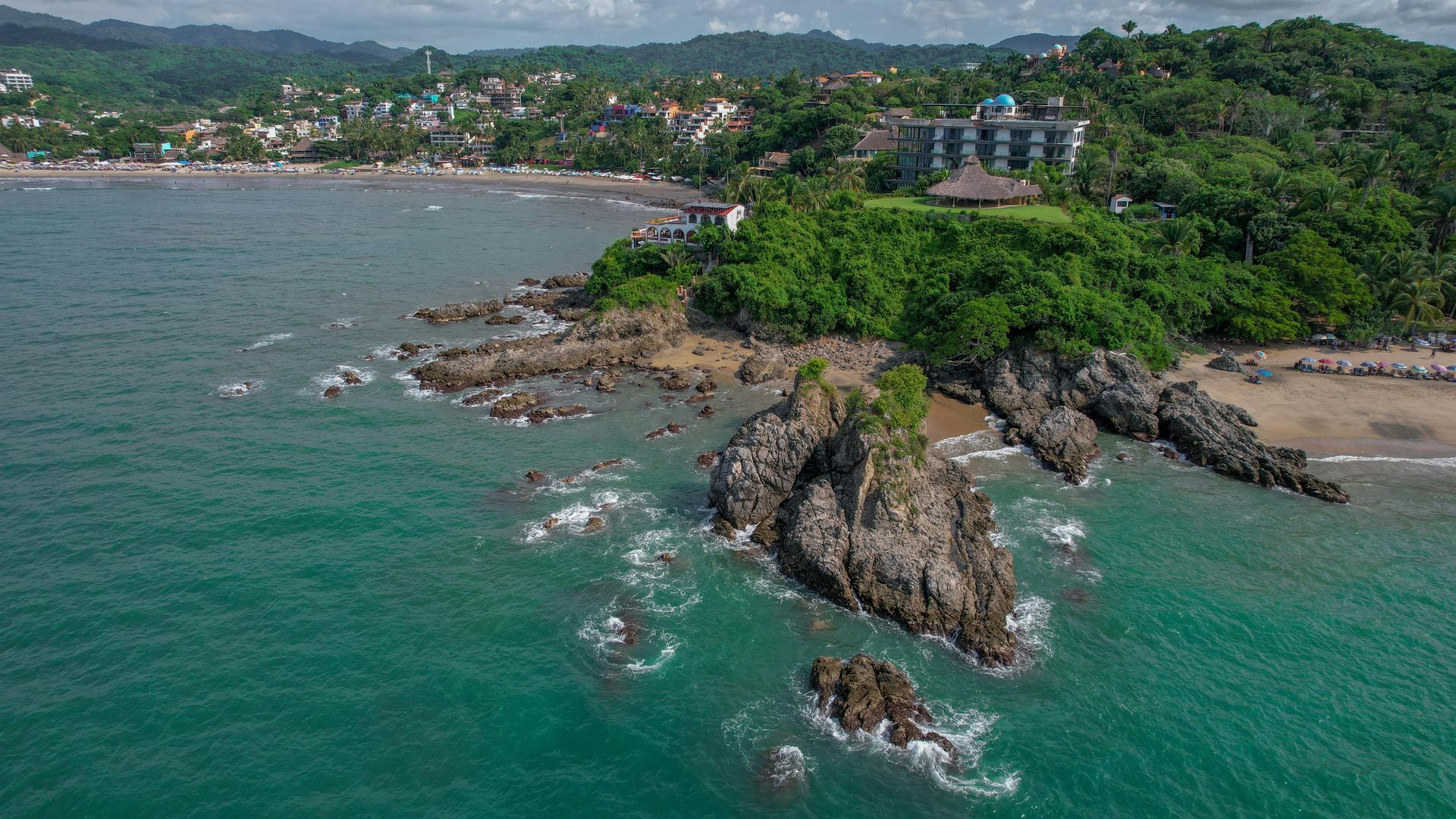 Rocky coastline meets turquoise ocean; green trees and buildings in the background.