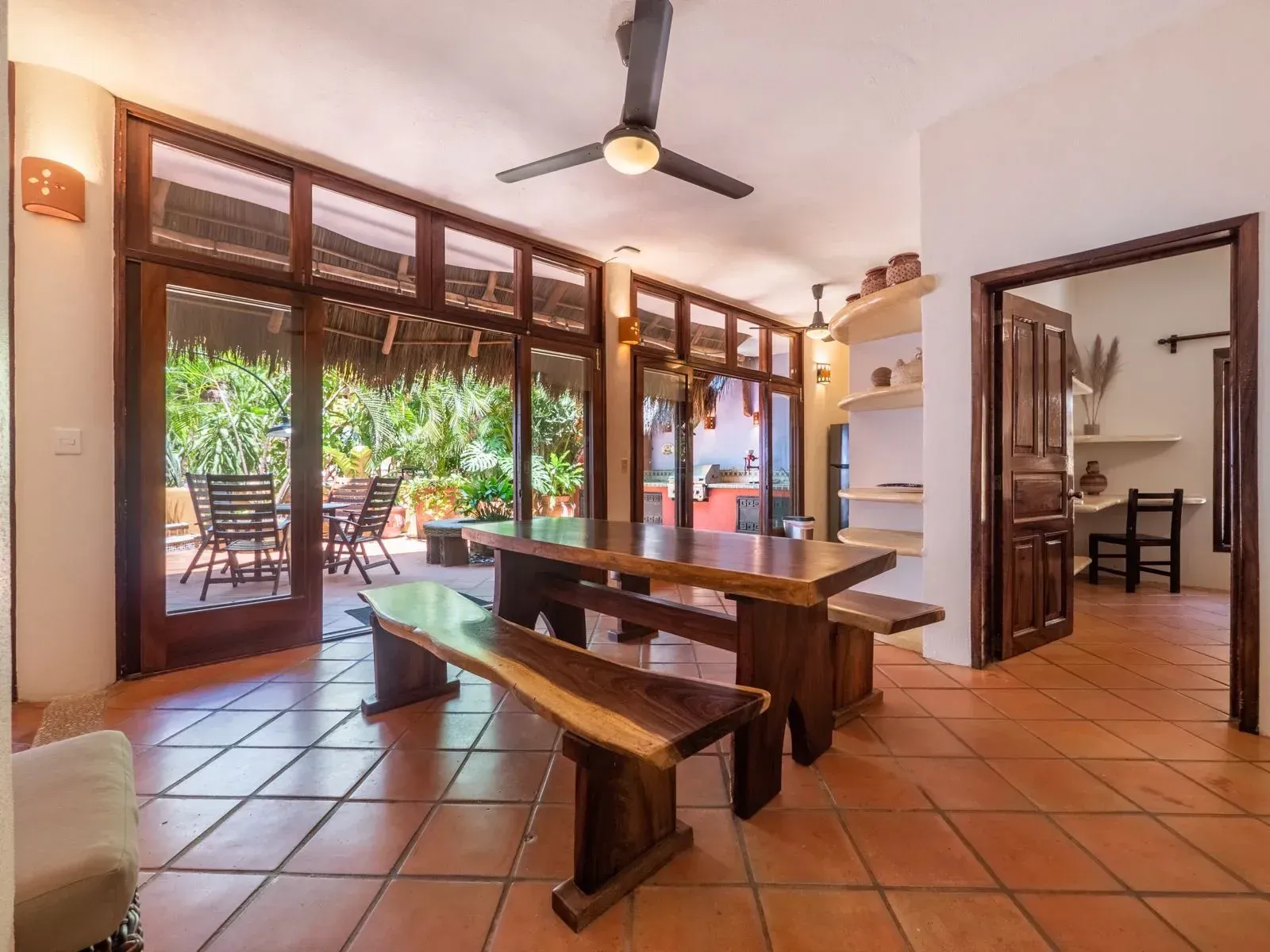 Dining room with wooden table, benches, and patio view, terracotta tiles and open doorway.