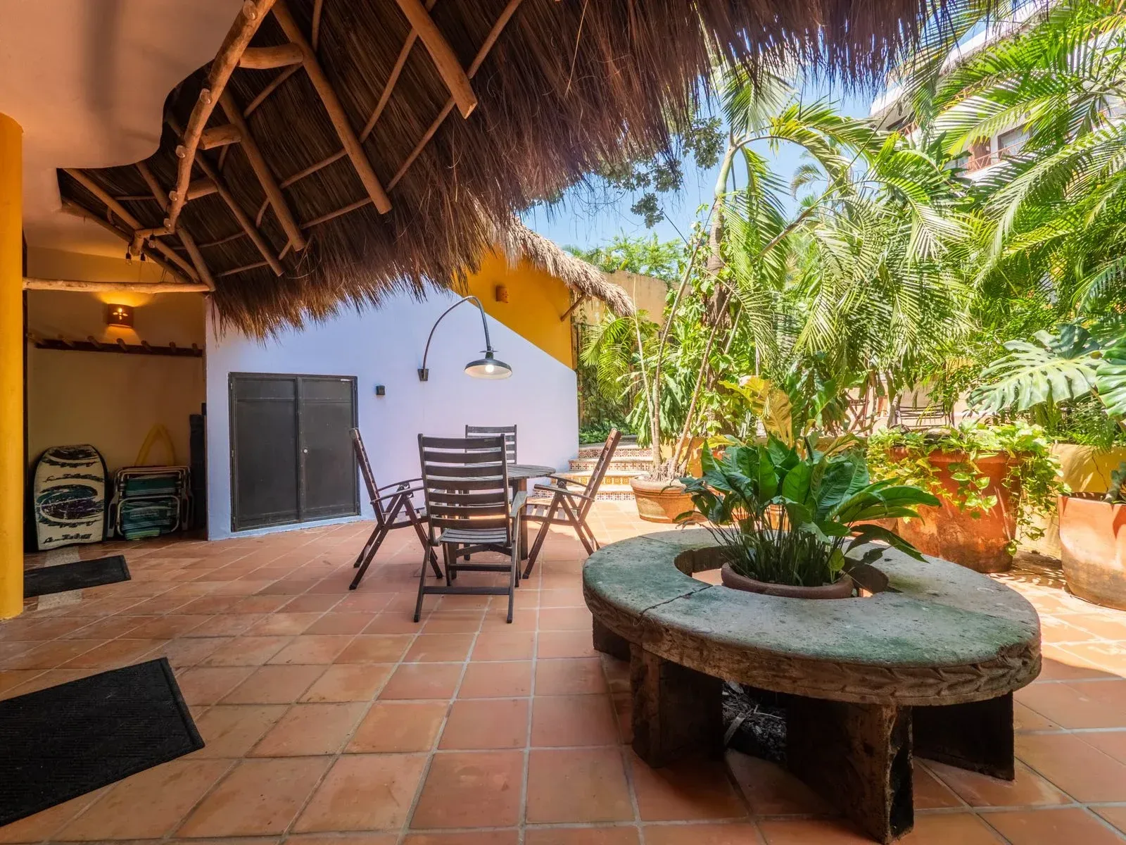 Patio with table, chairs, stone planter, and thatched roof in a tropical setting.