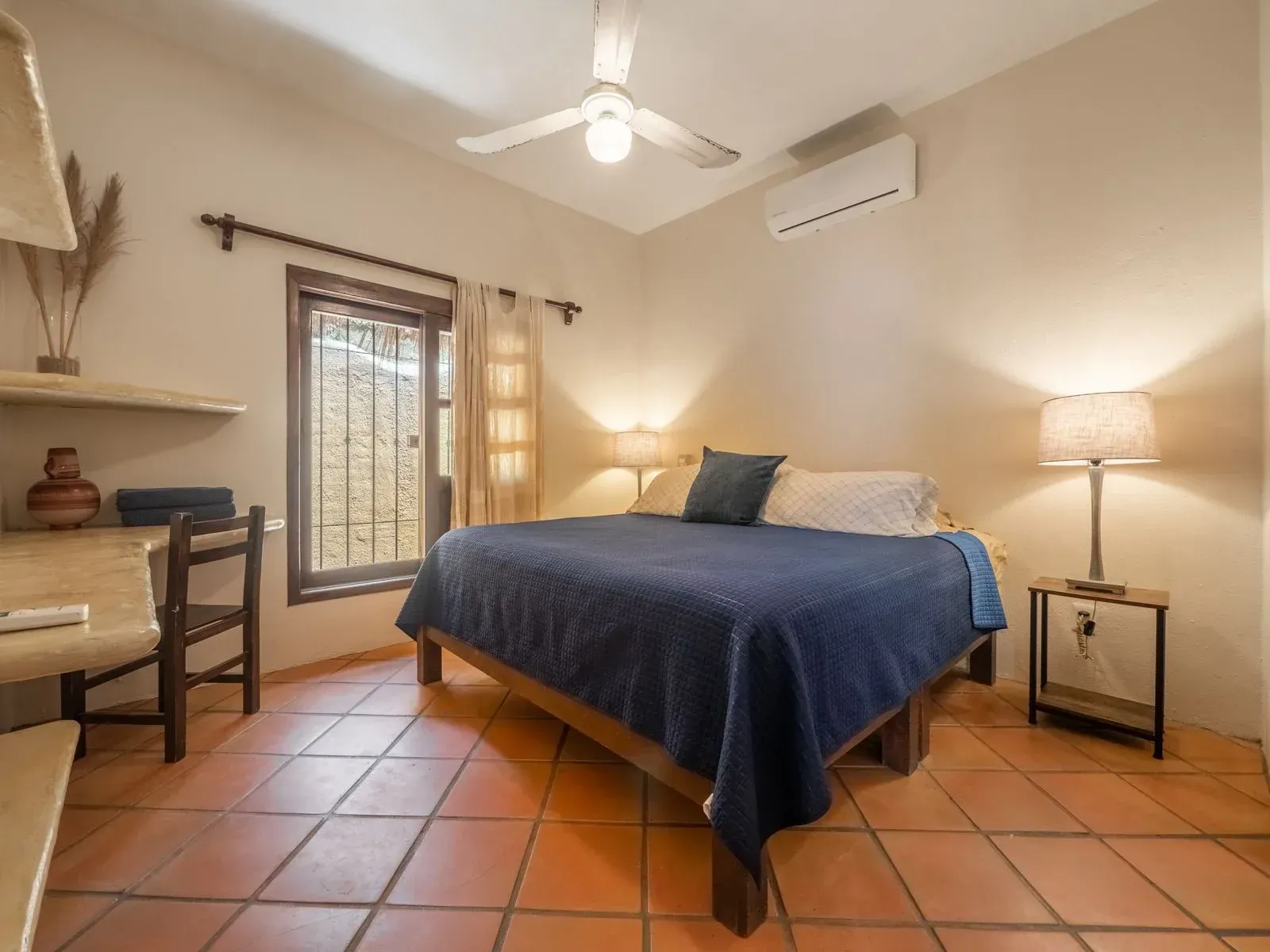 Bedroom with a bed, desk, window, and terracotta tile floor. Warm lighting and a ceiling fan.