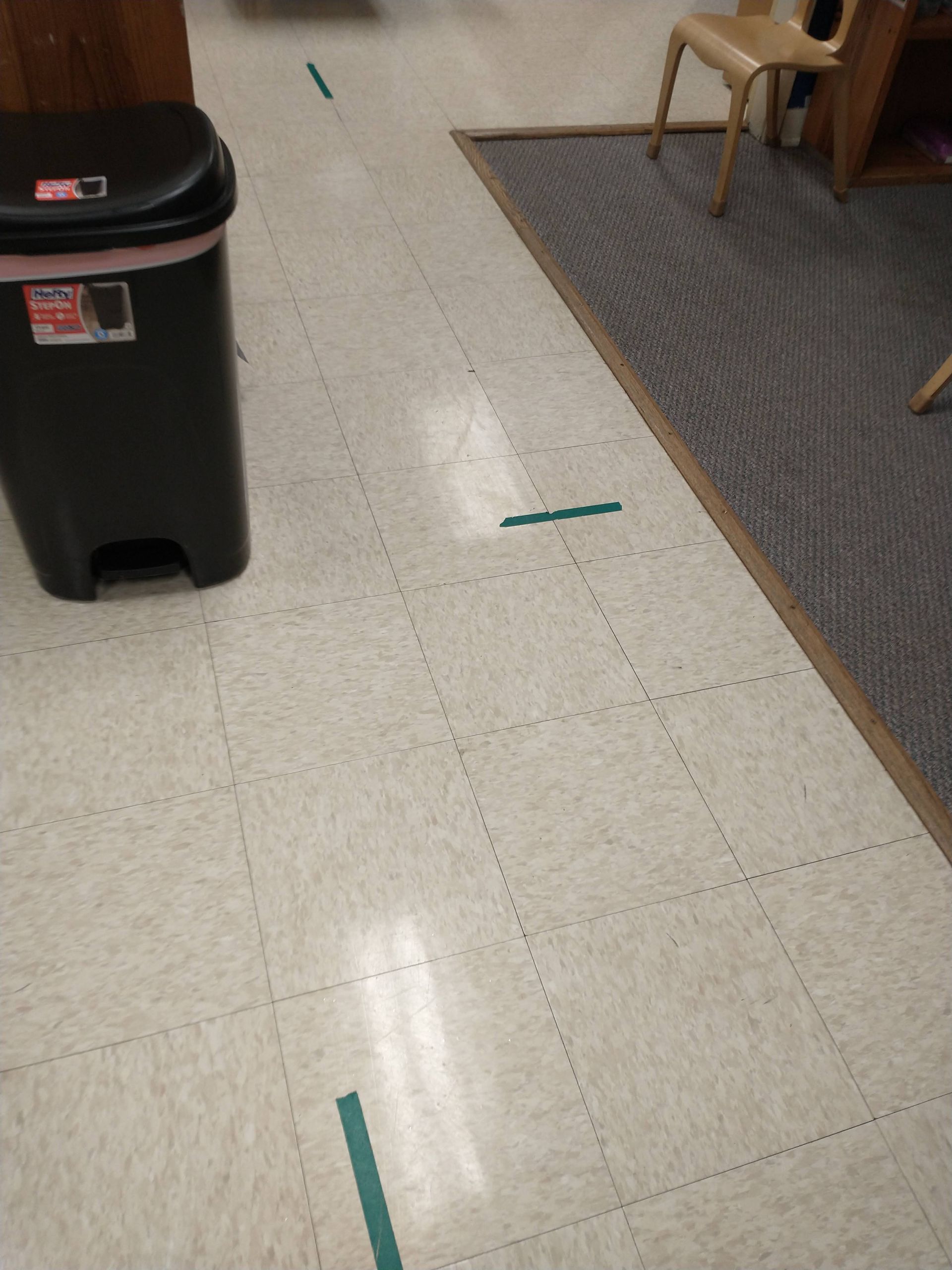 Hallway floor with teal tape markers, a black trash bin, and a chair on a carpeted mat