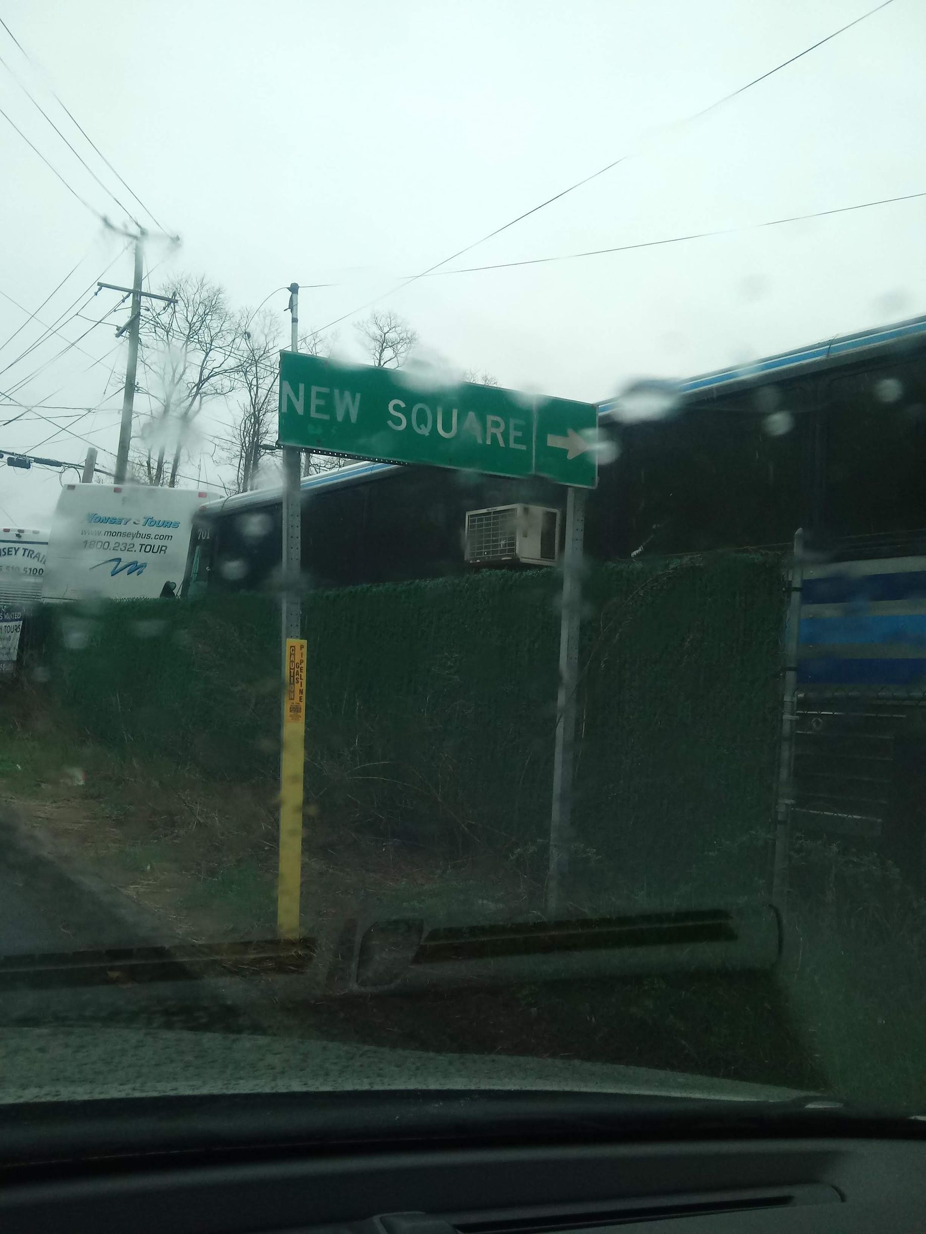 Rainy street view of a green “NEW SQUARE” sign by a fenced building, seen through a car windshield