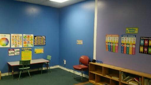 Empty classroom corner with blue walls, small tables, chairs, cubbies, and colorful learning posters.