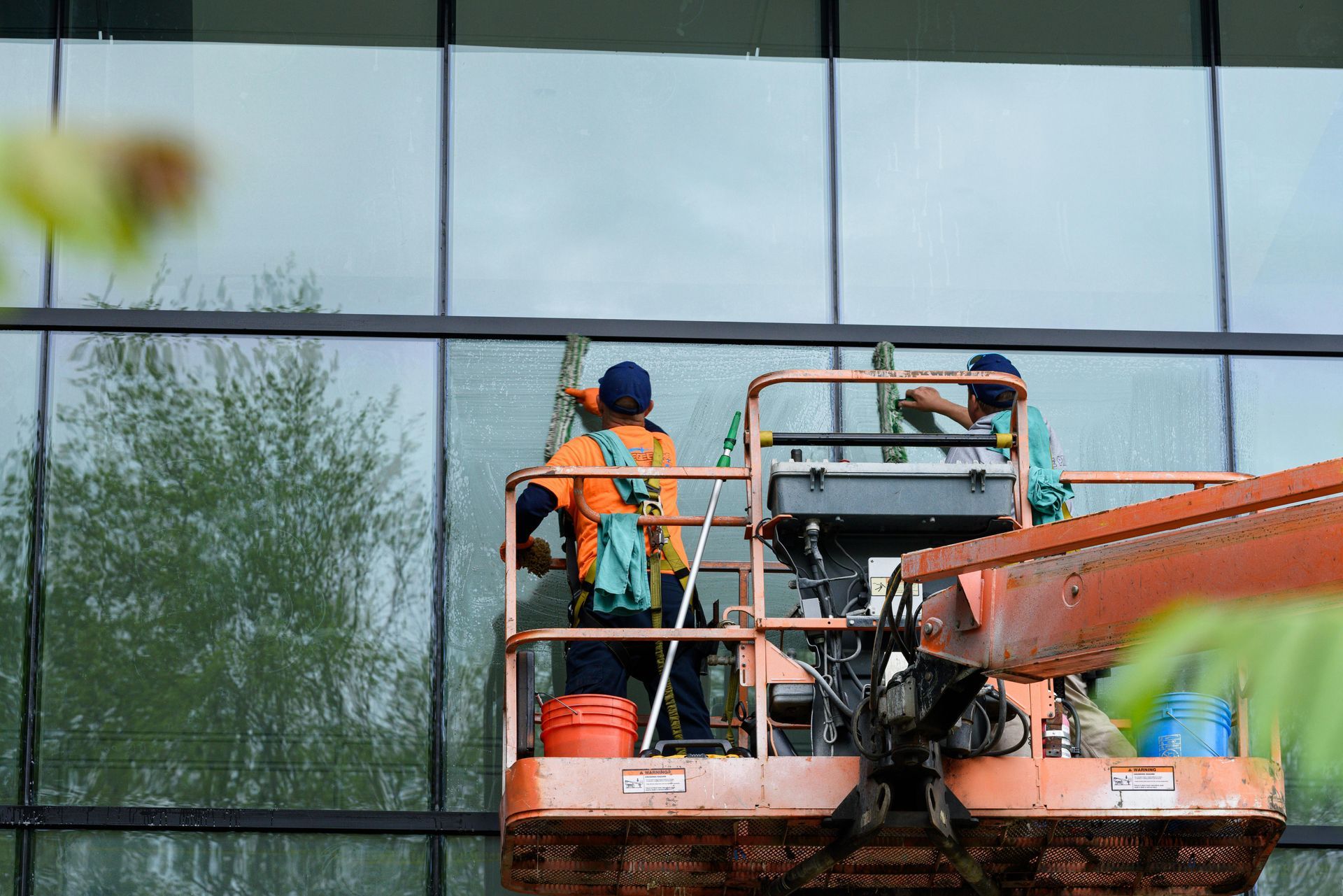 Workers on an orange lift cleaning a glass building facade with water spray.