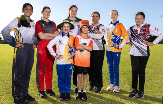 Group of Girls from Ascot Junior Girls Cricket teams in colorful sports uniforms standing on a grassy field, posing together and smiling.