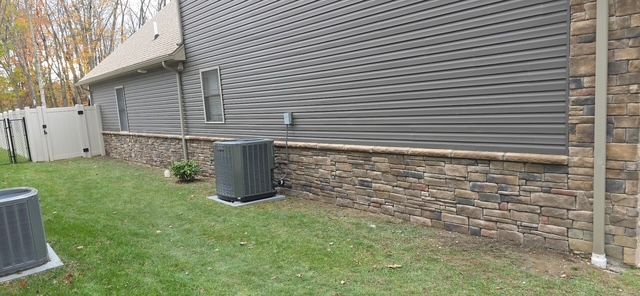 Side of a house with gray siding and stone veneer base, air conditioning unit in yard.
