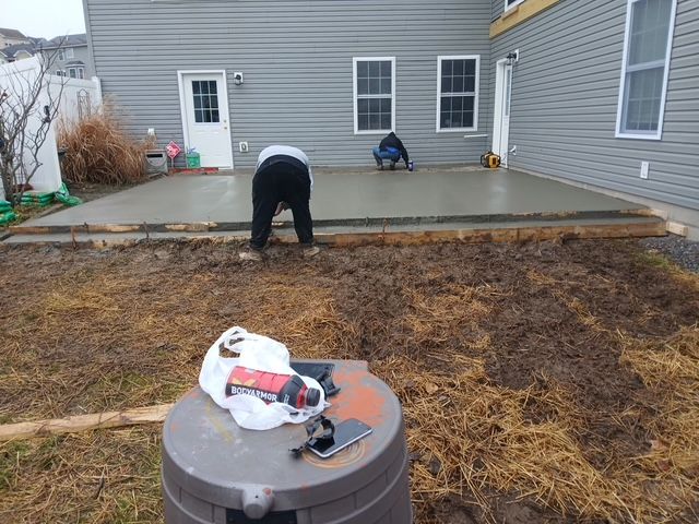 Workers leveling concrete patio in backyard of a house, with surrounding mud and tools.