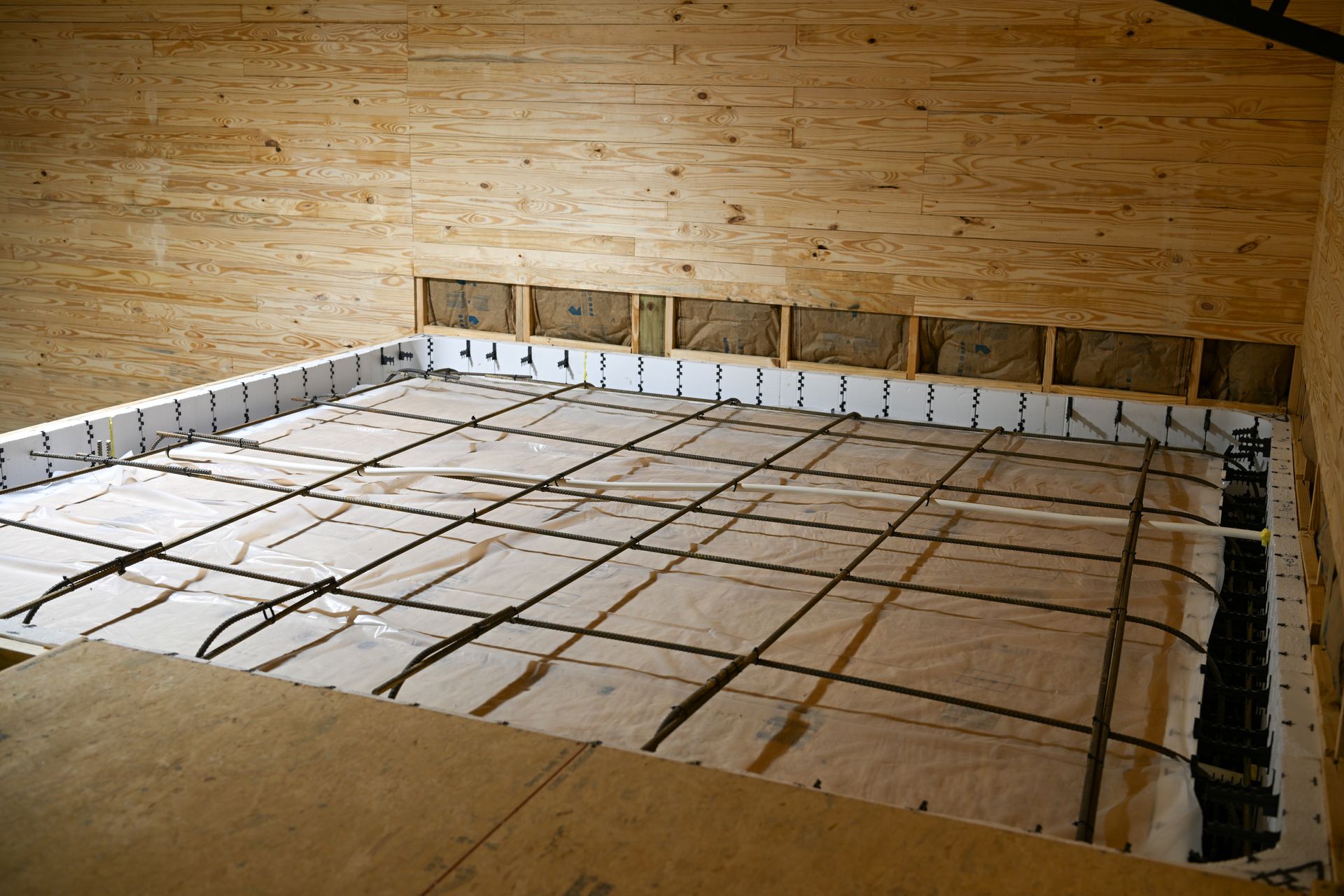 A view of a concrete floor foundation being prepared with a rebar grid, white vapor barrier, and foam insulation boards.