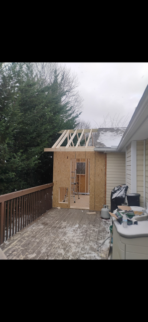 A wooden structure in progress on a deck, with framing and sheathing visible. Trees and a house exterior are in the background.