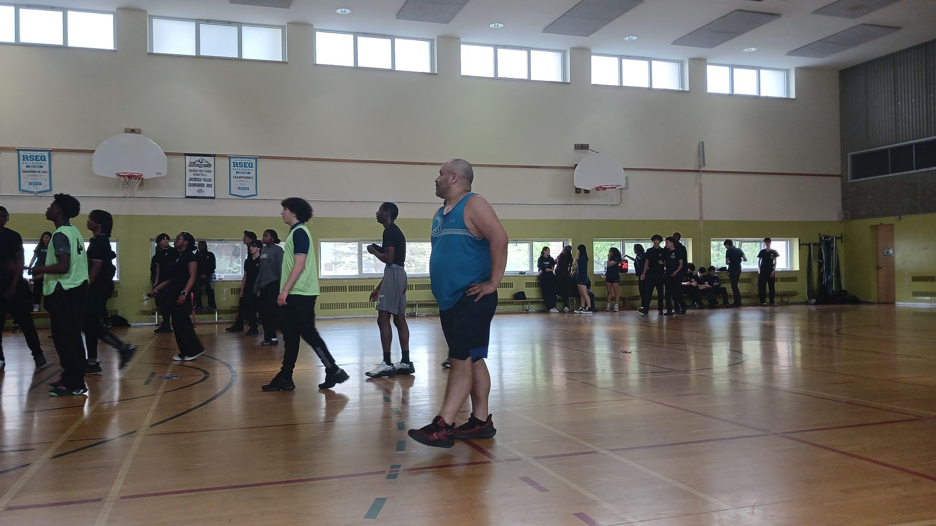 Gymnasium with people standing and walking. A man in a tank top stands in the foreground, facing away from the camera.