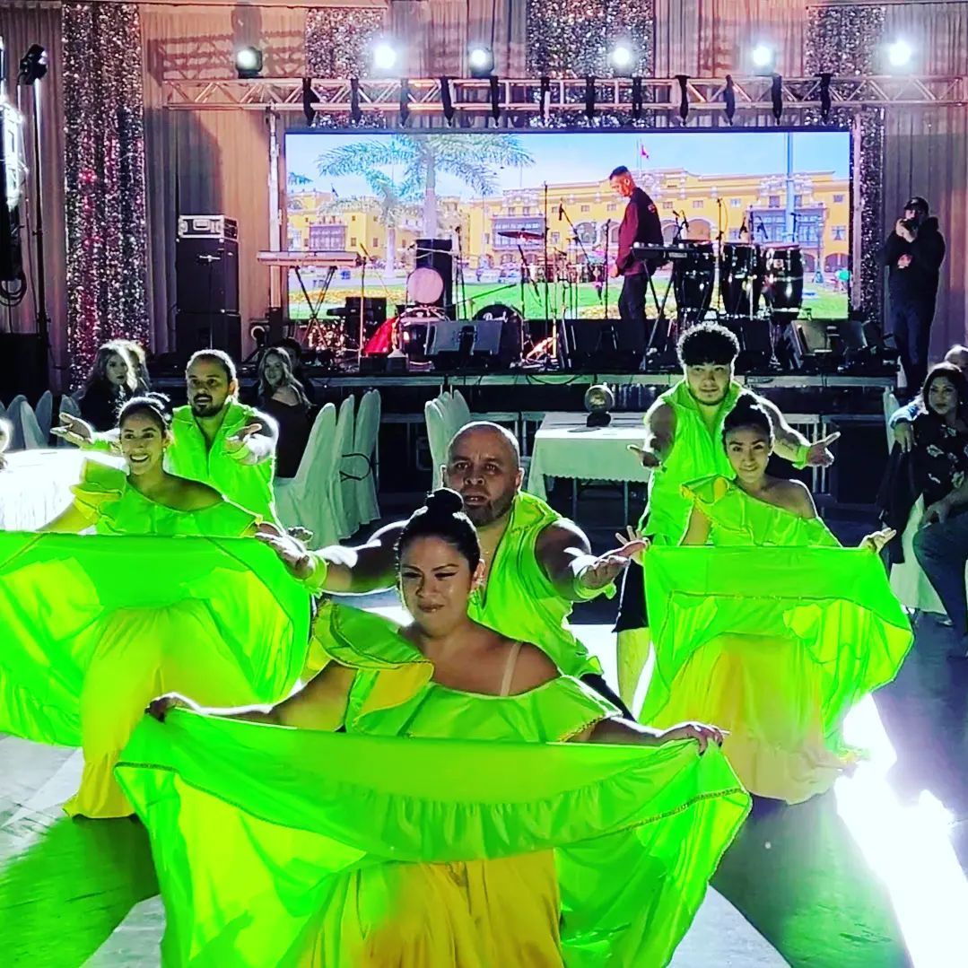 A group of dancers in bright green and yellow outfits perform in front of a stage with a band.